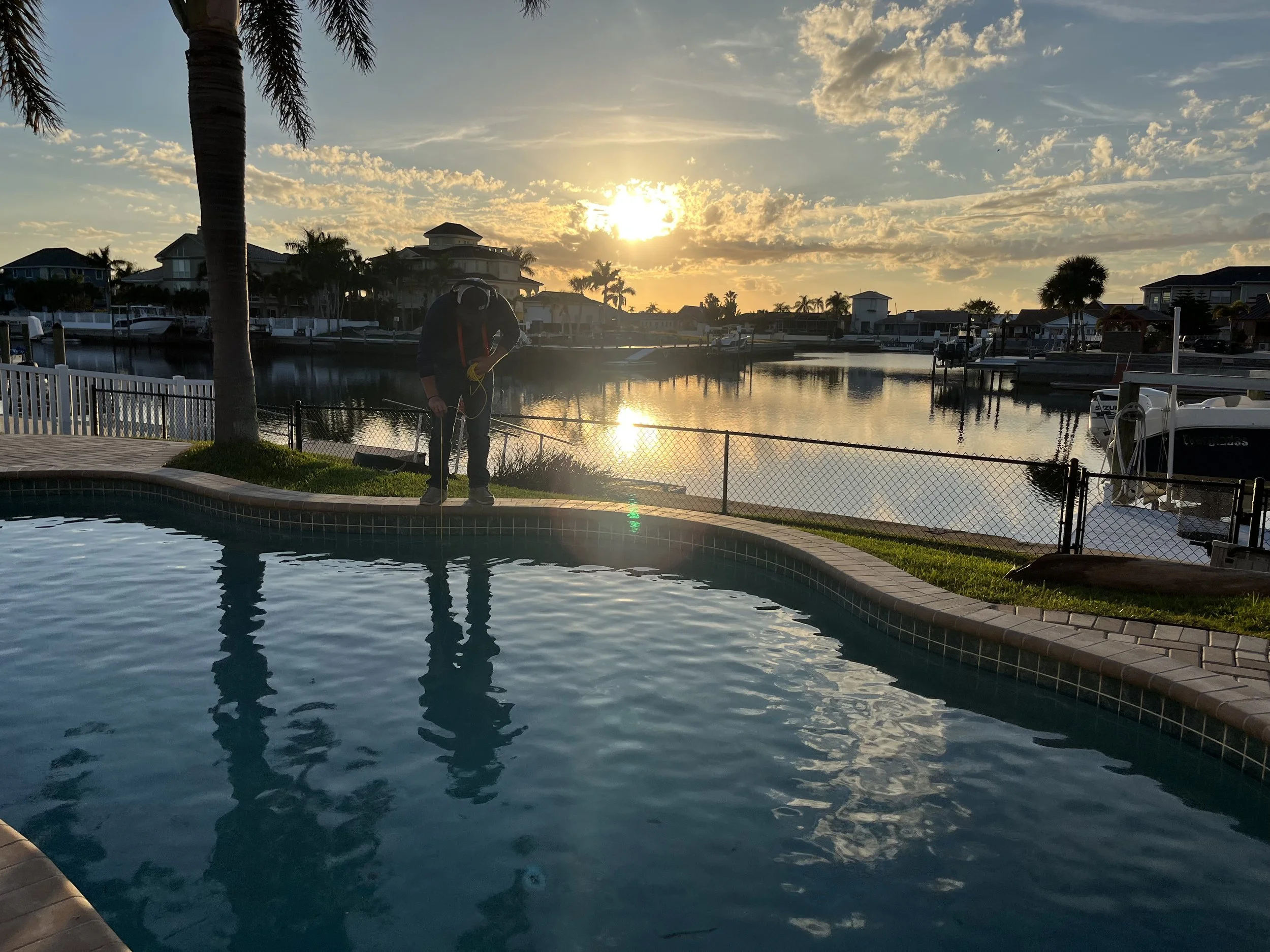A person wearing scuba gear stands on the edge of a swimming pool near a waterfront property during sunset, with houses and boats visible across the water.