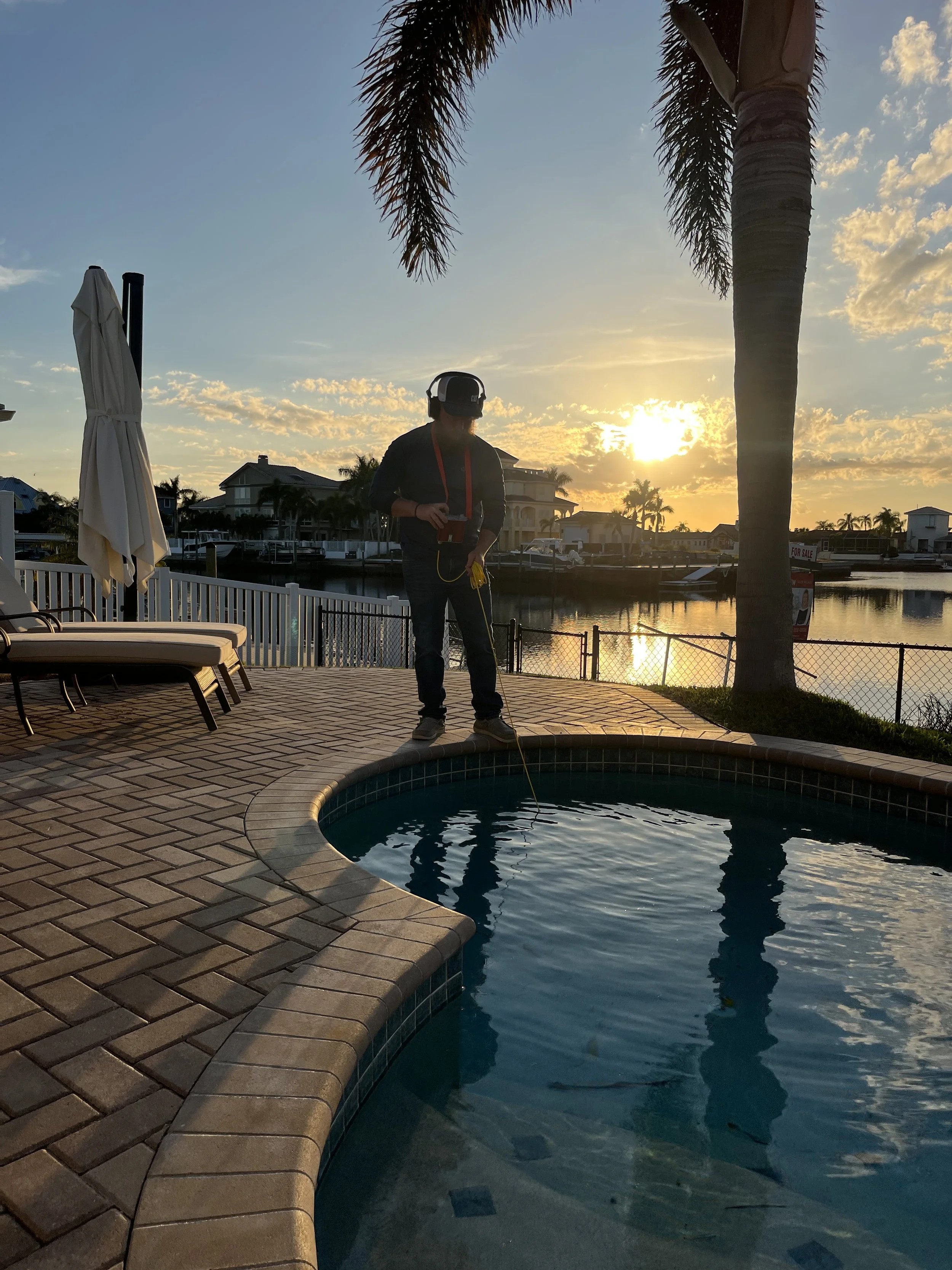 Person standing beside a backyard swimming pool at sunset, wearing headphones and holding a metal detector, with a waterfront view of houses and palm trees in the background.