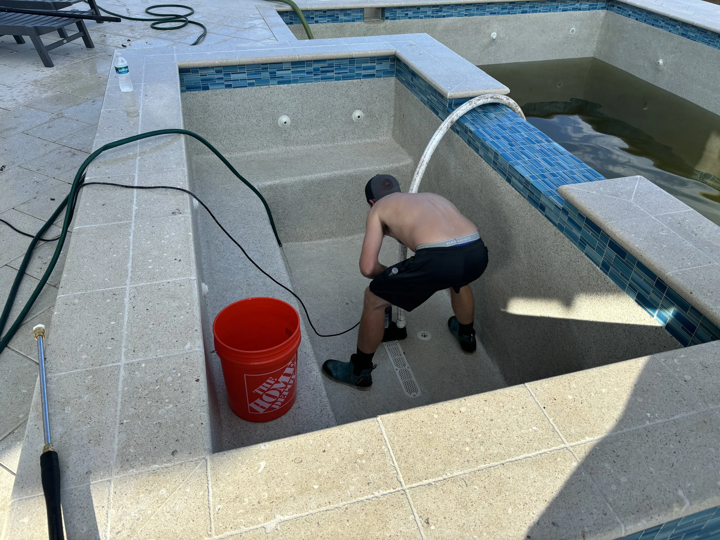 A shirtless young man wearing black shorts and a black cap is cleaning or repairing an empty hot tub or pool with beige and blue tiles, surrounded by patio tiles. A red bucket and cleaning tools are nearby.