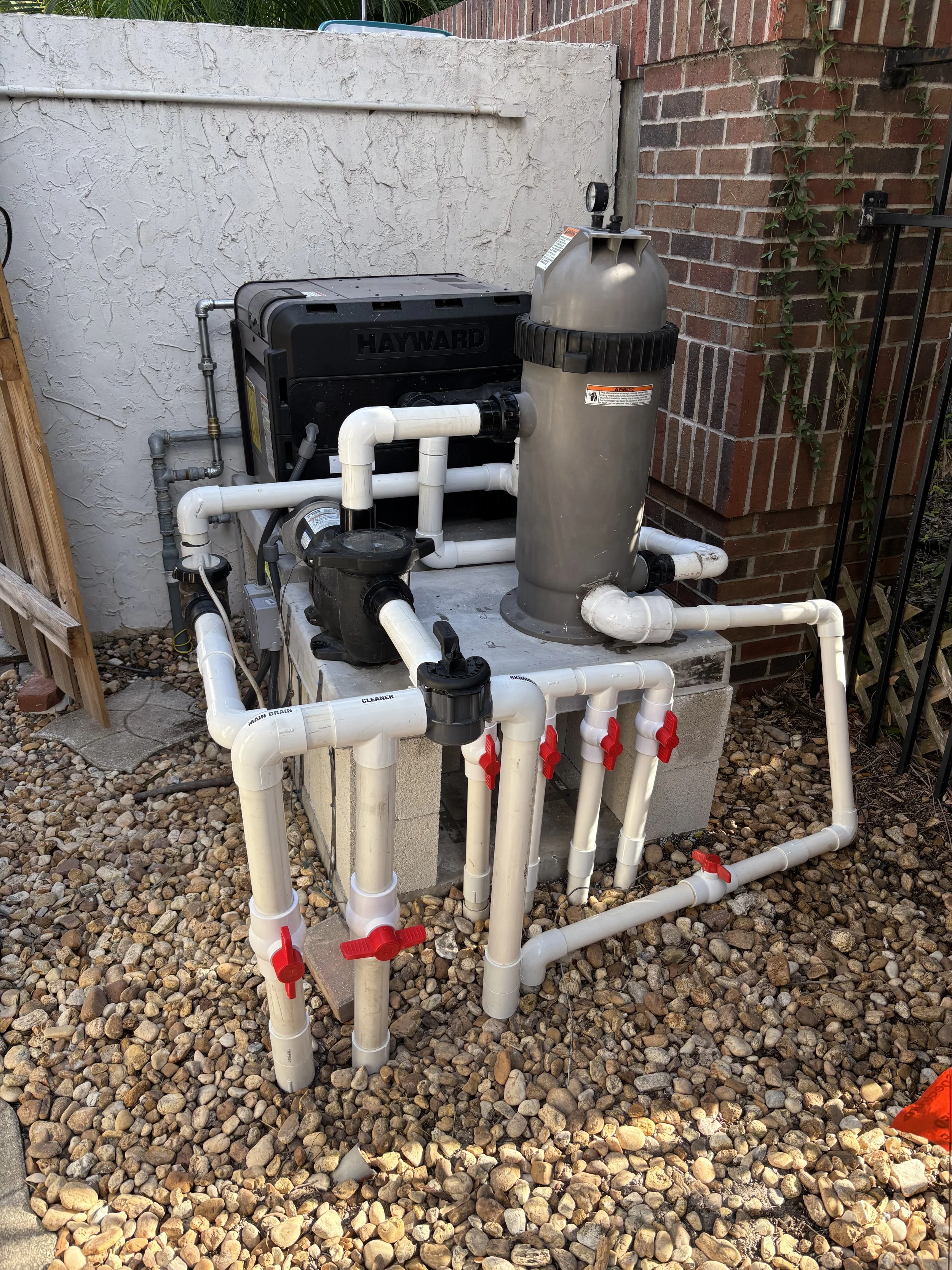 Pool filtration system with white PVC pipes, a sand filter, and a black Hayward pump, installed outdoors on a concrete platform with gravel ground, next to a brick wall and a white stucco wall.