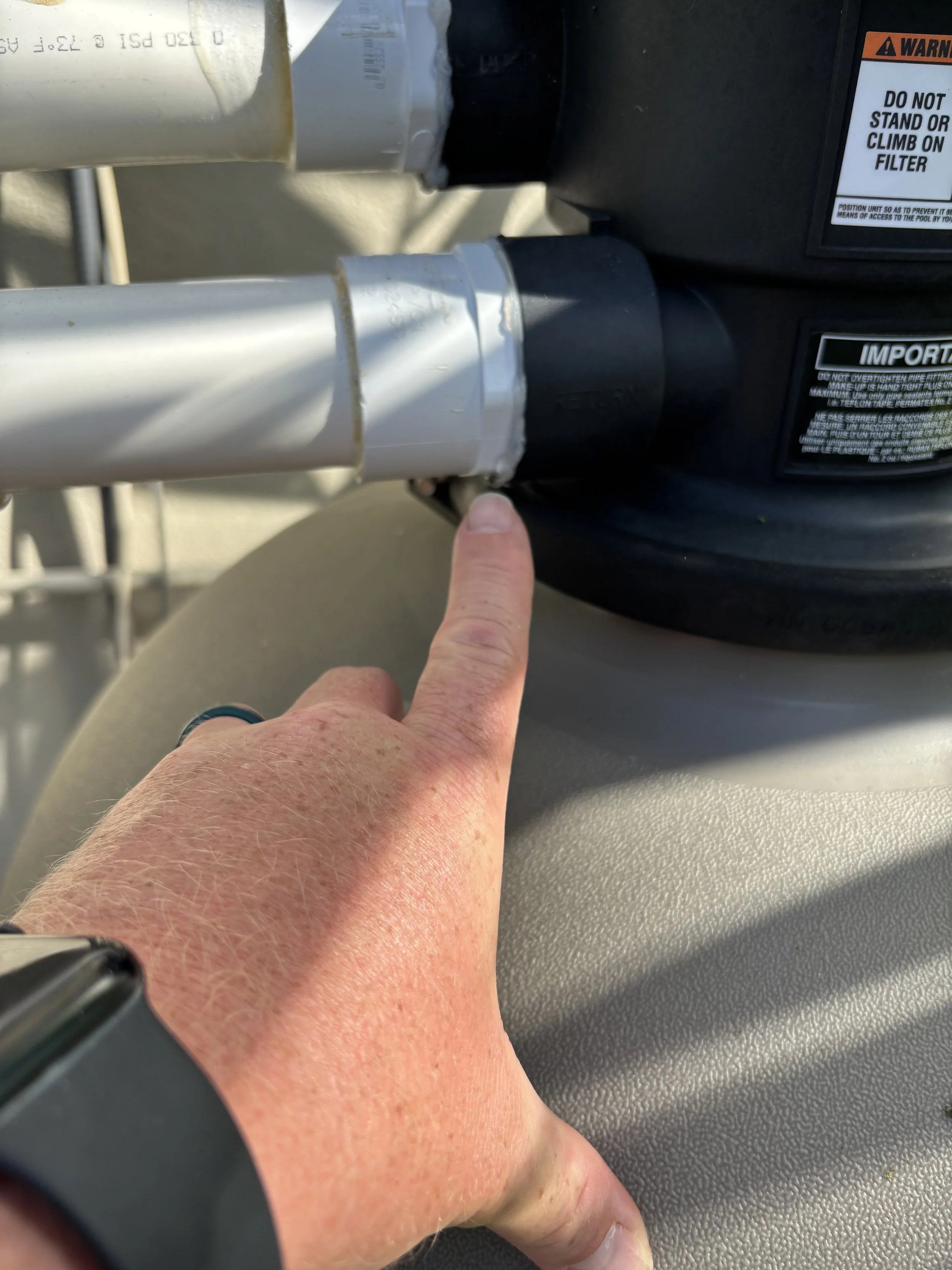 A person's hand pointing at a valve connection on a pool pump filter, with pipes attached.