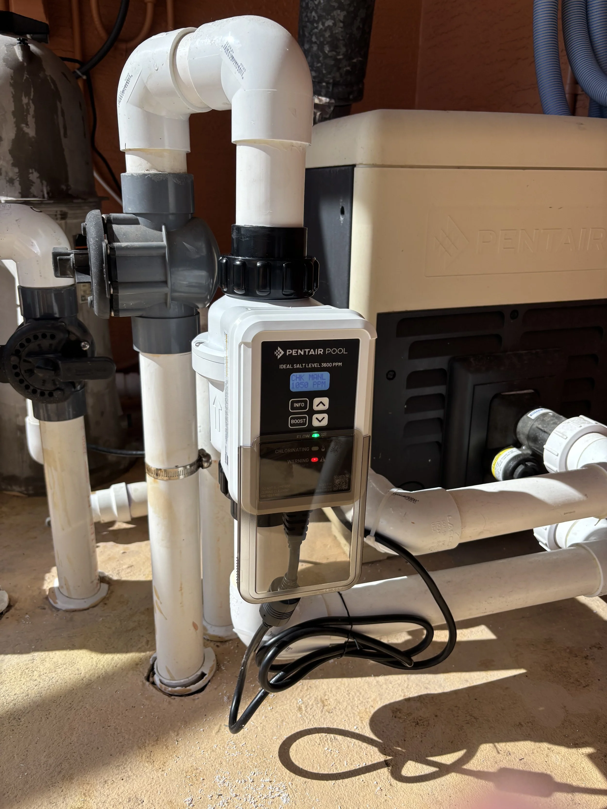 Pool salt chlorinator system with white and black components, pipes, and control unit, set up outdoors on a concrete surface near a Pentair pool equipment unit.