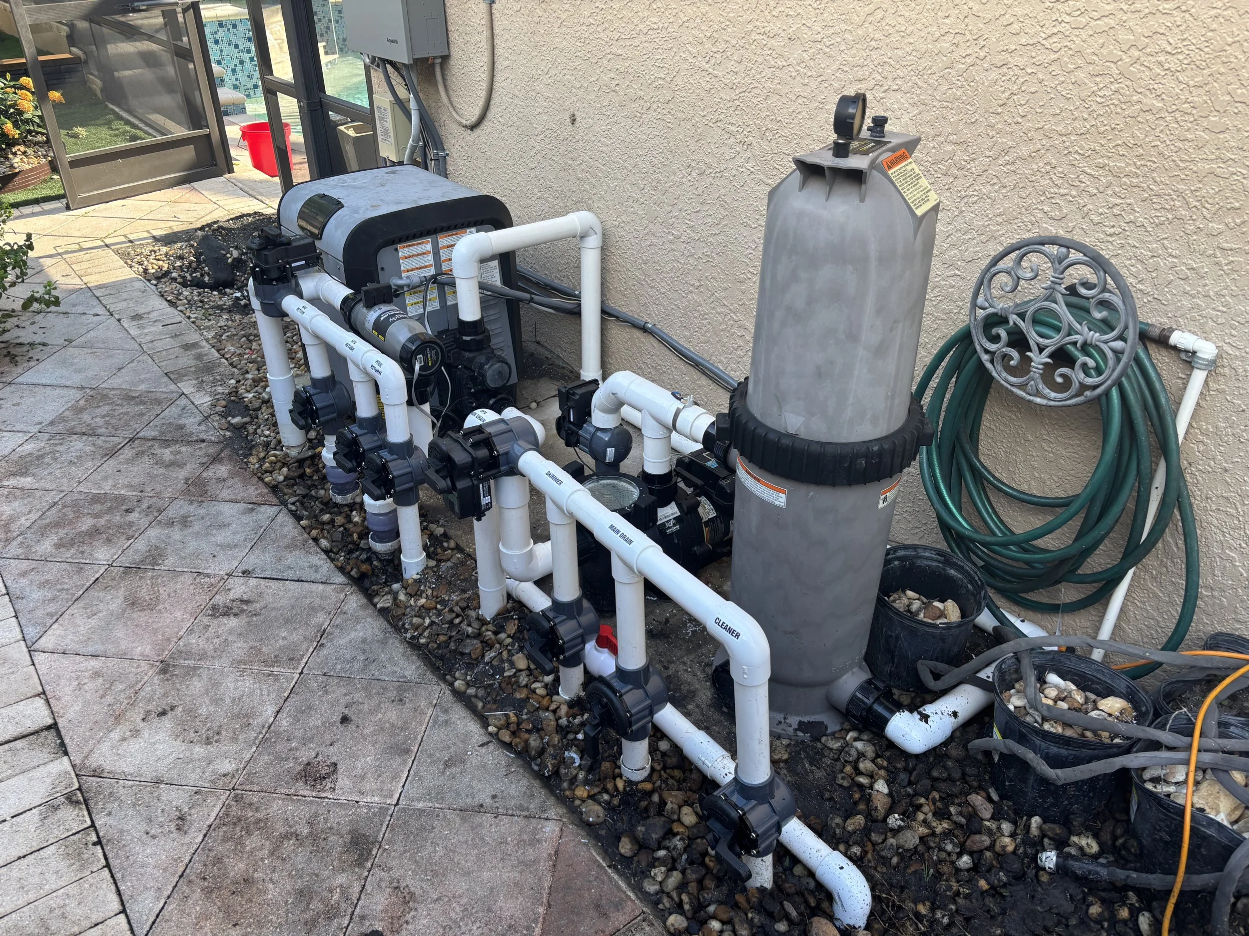 Pool equipment setup with filter, pump, and piping next to a beige stucco wall, with a garden hose and small buckets on the ground.