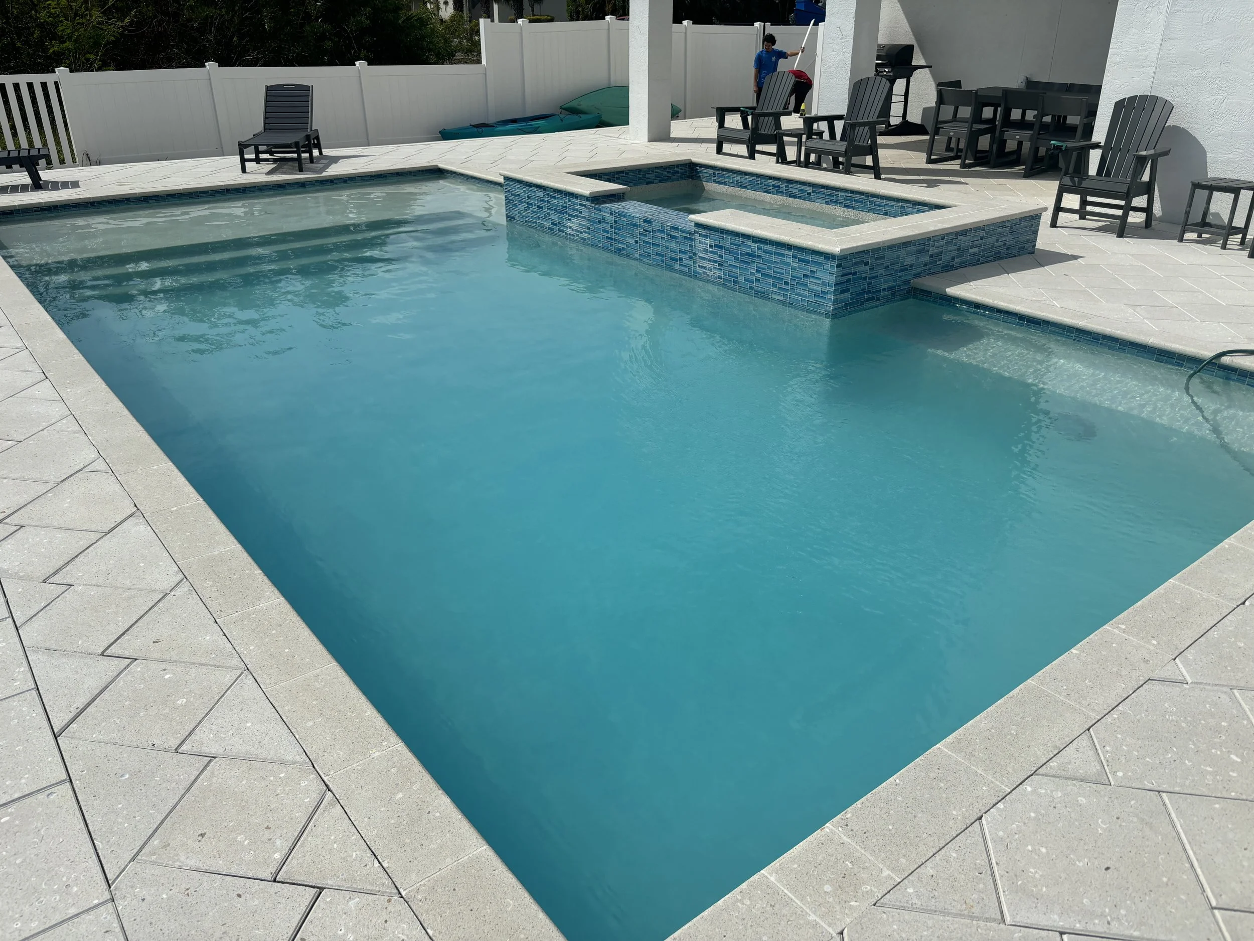 Residential backyard with a swimming pool, hot tub, and patio seating area. There are chairs, a table, and a grill. Two children are playing near the fence.