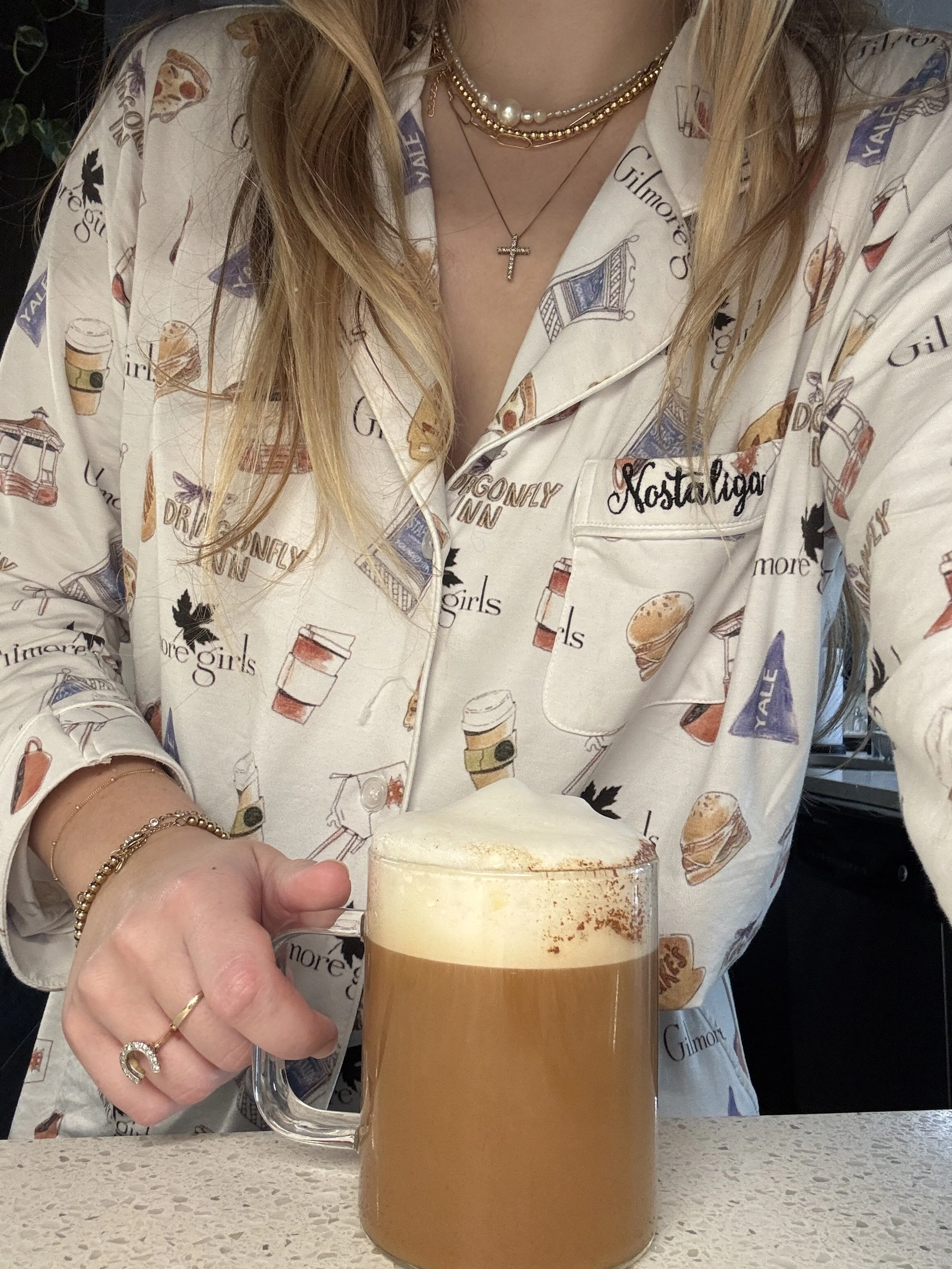A woman wearing a white pajama top with various food and coffee illustrations and the words "Gilmore Girls" and "Nostalgia" embroidered on it, holding a glass mug of coffee with foam on top. She has blonde hair, layered necklaces including a cross, and a bracelet and ring on her left hand.