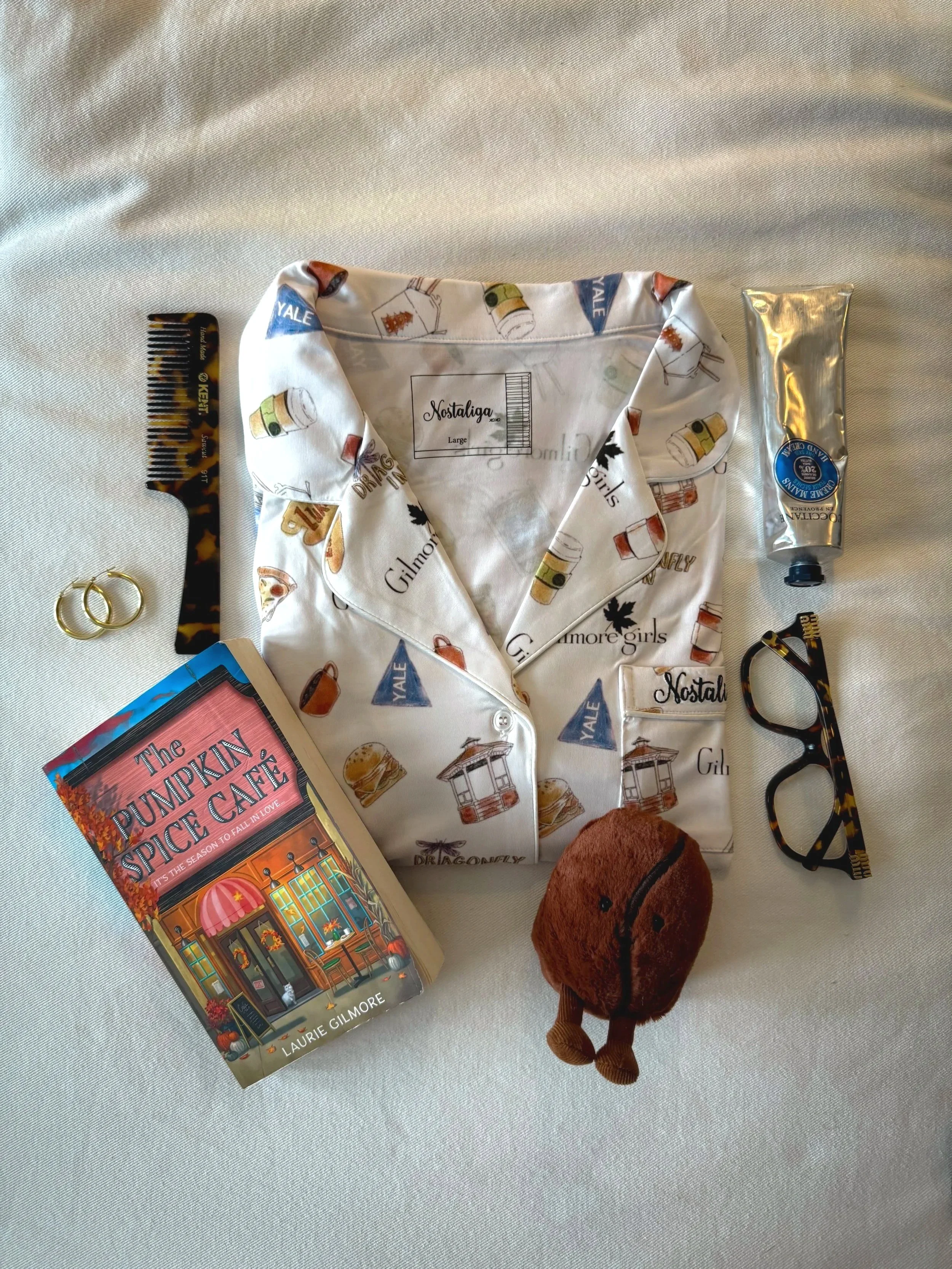 A collection of personal items on a white surface including a book titled 'The Pumpkin Spice Cafe', a toy plush of a brown animal, a pair of tortoise-shell glasses, a tube of hand cream, a black and tortoise-shell patterned hair clip, gold hoop earrings, a packet of hair color, and a white button-up shirt with a pattern of drinks and popcorn and the brand name Gilmour Girls.
