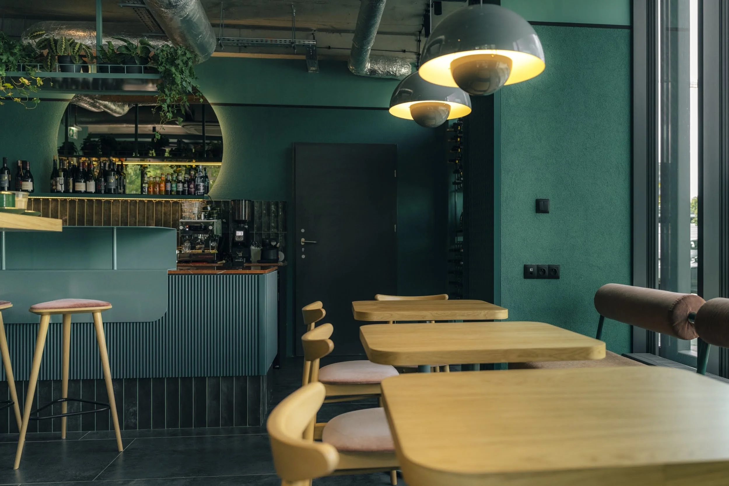 Interior of a modern cafe with green walls, wooden tables and chairs, black hanging lights, and a bar area with bottles and coffee equipment.