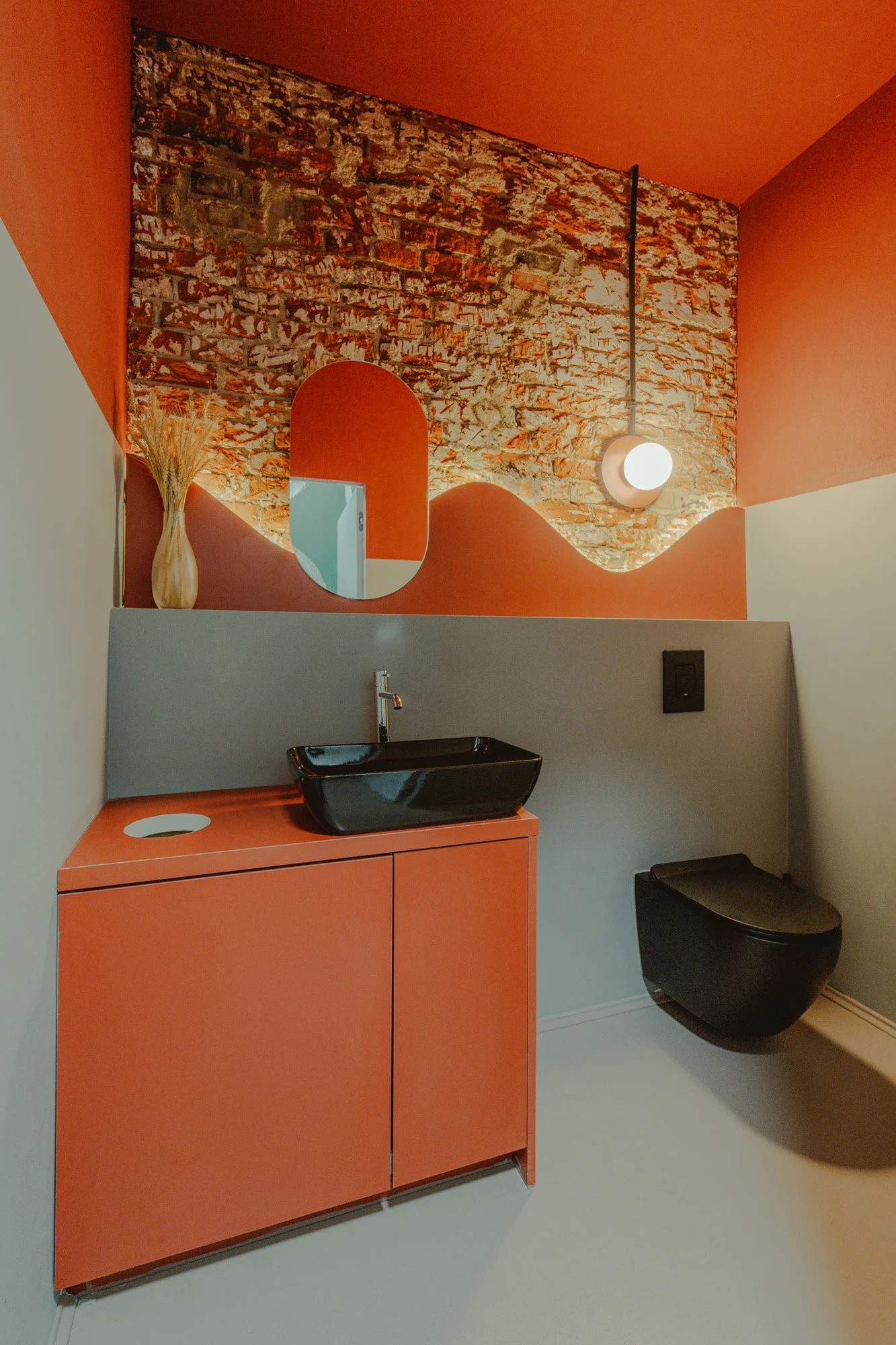 Modern bathroom with orange and gray color scheme, exposed brick wall, black sink and toilet, decorative vase, round mirror, and spherical wall light.