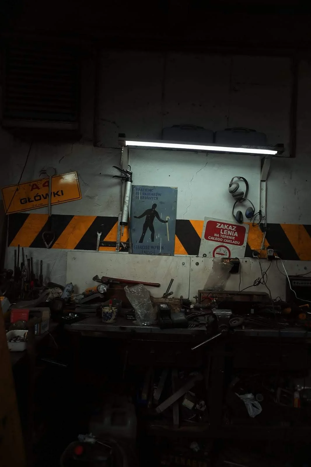 A cluttered workshop with a workbench, tools, and signs on the wall, including a yellow sign with black text and a red sign with white text. A fluorescent light illuminates the wall and some objects are hanging, such as headphones and a screwdriver.