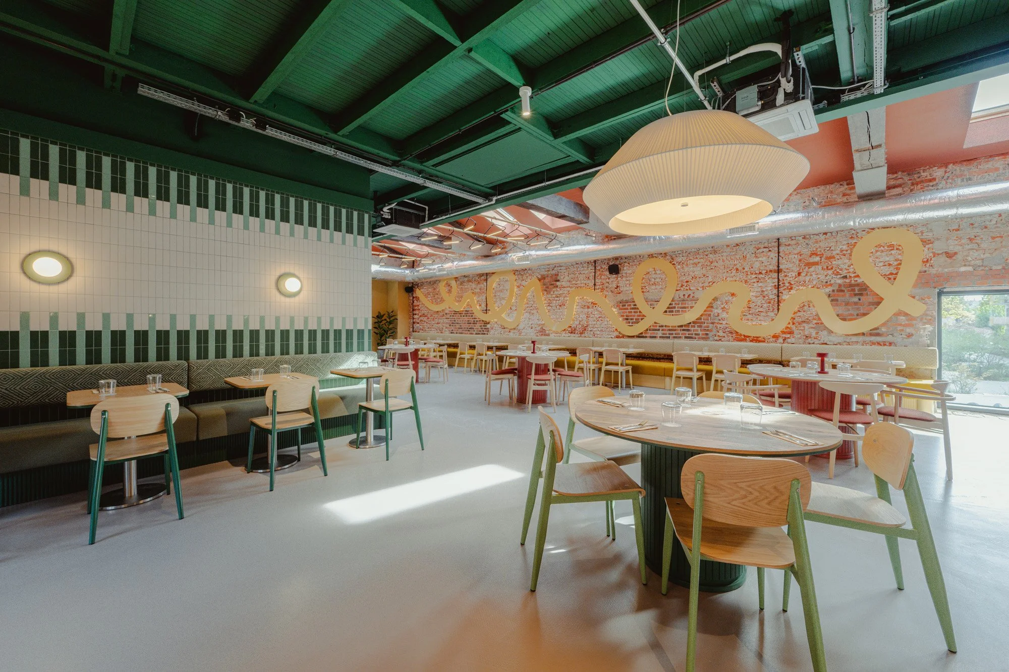 Empty modern restaurant with green and natural wood furniture, a brick wall with yellow abstract art, and a large cream-colored hanging light.