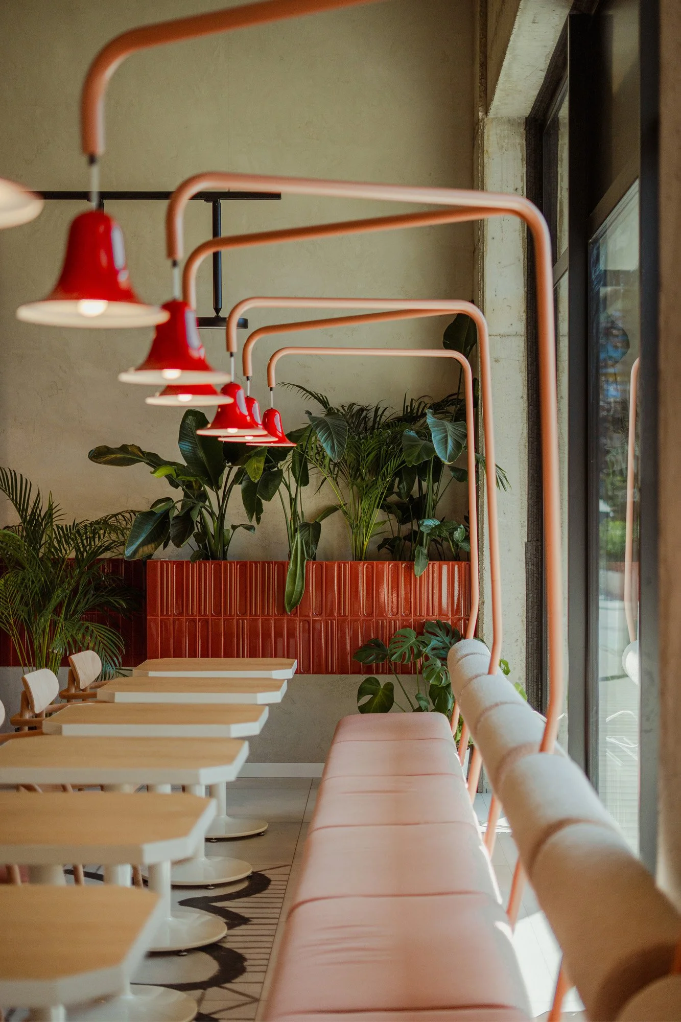 Interior of a cafe with pink pendant lights, beige seating, white tables, and green plants in a red planter box along a window.