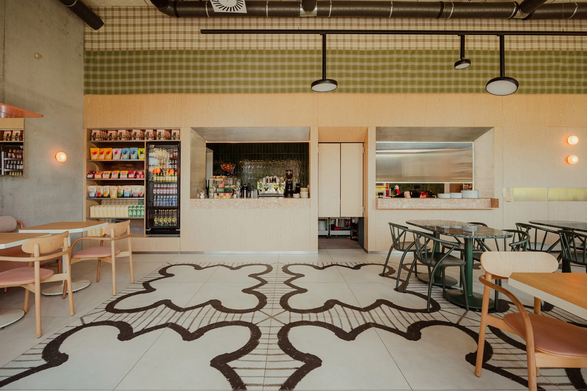 Interior of a modern café with tables and chairs, a patterned floor, and a counter area with a coffee machine and shelves stocked with snacks and drinks.