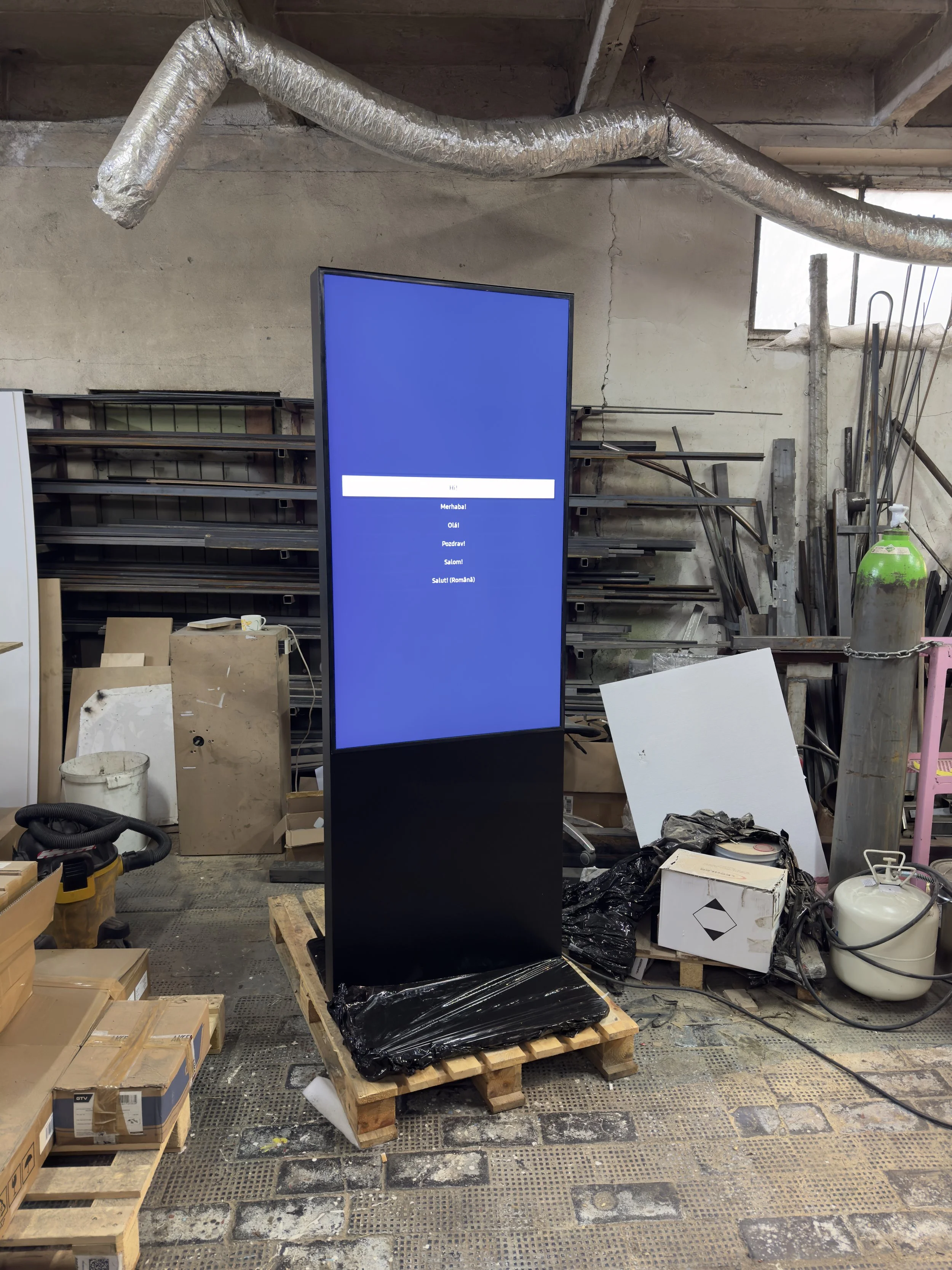 A digital sign or kiosk with a blue screen displaying text options in different languages, placed on a wooden pallet in a workshop or storage area with tools, boxes, and construction materials around.