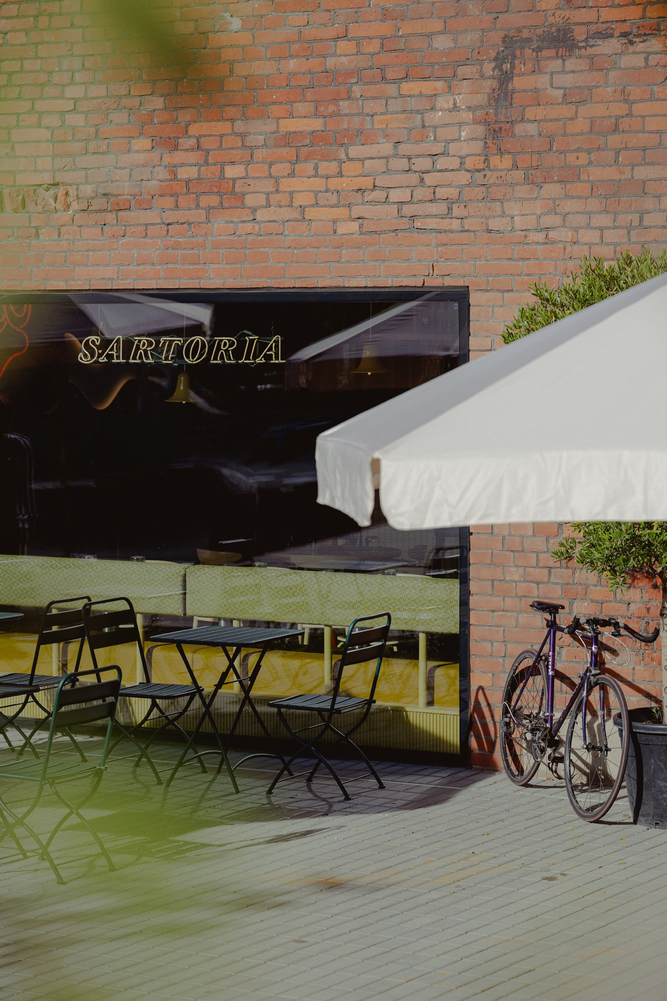 Outdoor patio with black chairs and tables, a white umbrella, a potted plant, a bicycle, and a storefront with a sign that reads 'SARTORIA' against a brick wall.