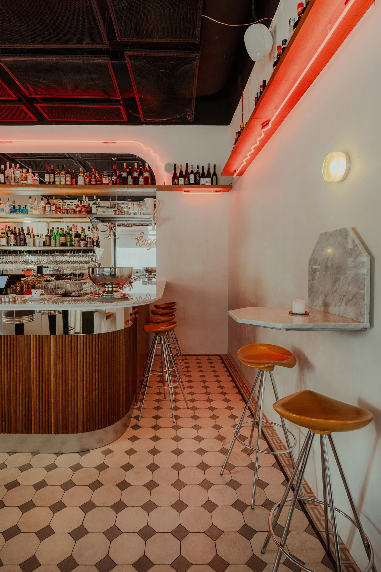 Interior view of a modern bar with a curved wooden counter, bar stools, shelves of various alcohol bottles, and a small table with stools along the wall, illuminated by warm lighting.