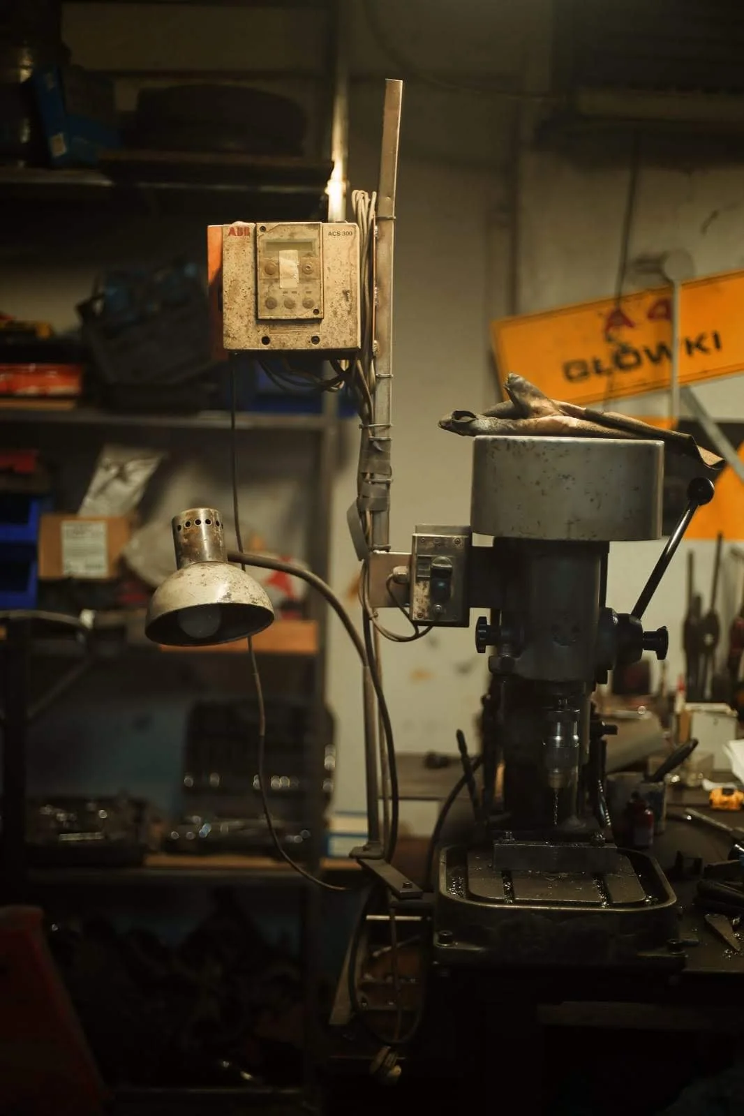A workshop containing a metal drill press, an industrial lamp, and various tools and boxes on shelves in the background.