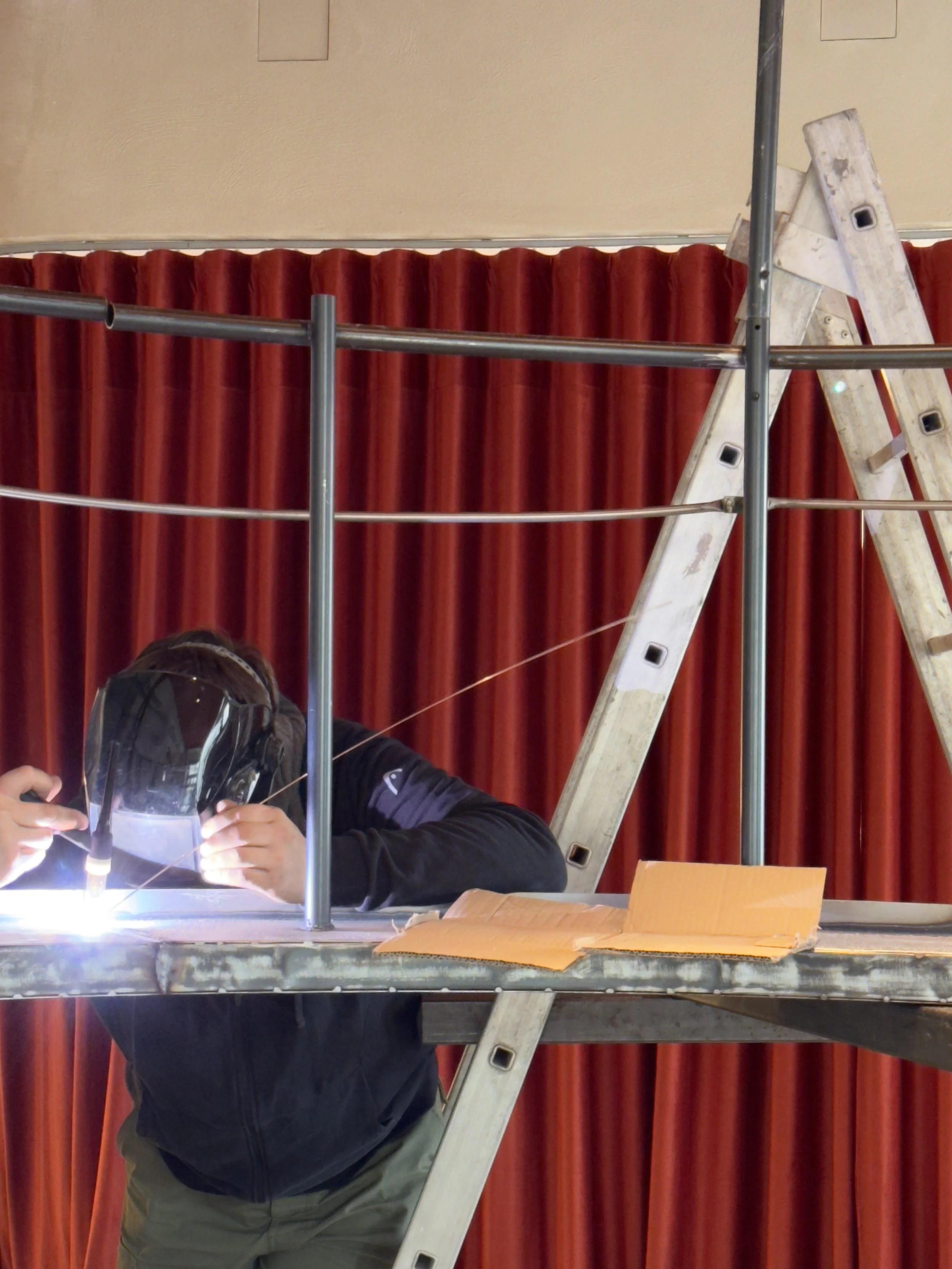 A person welding on a metal framework, using a helmet for safety, with a ladder and a red curtain in the background.