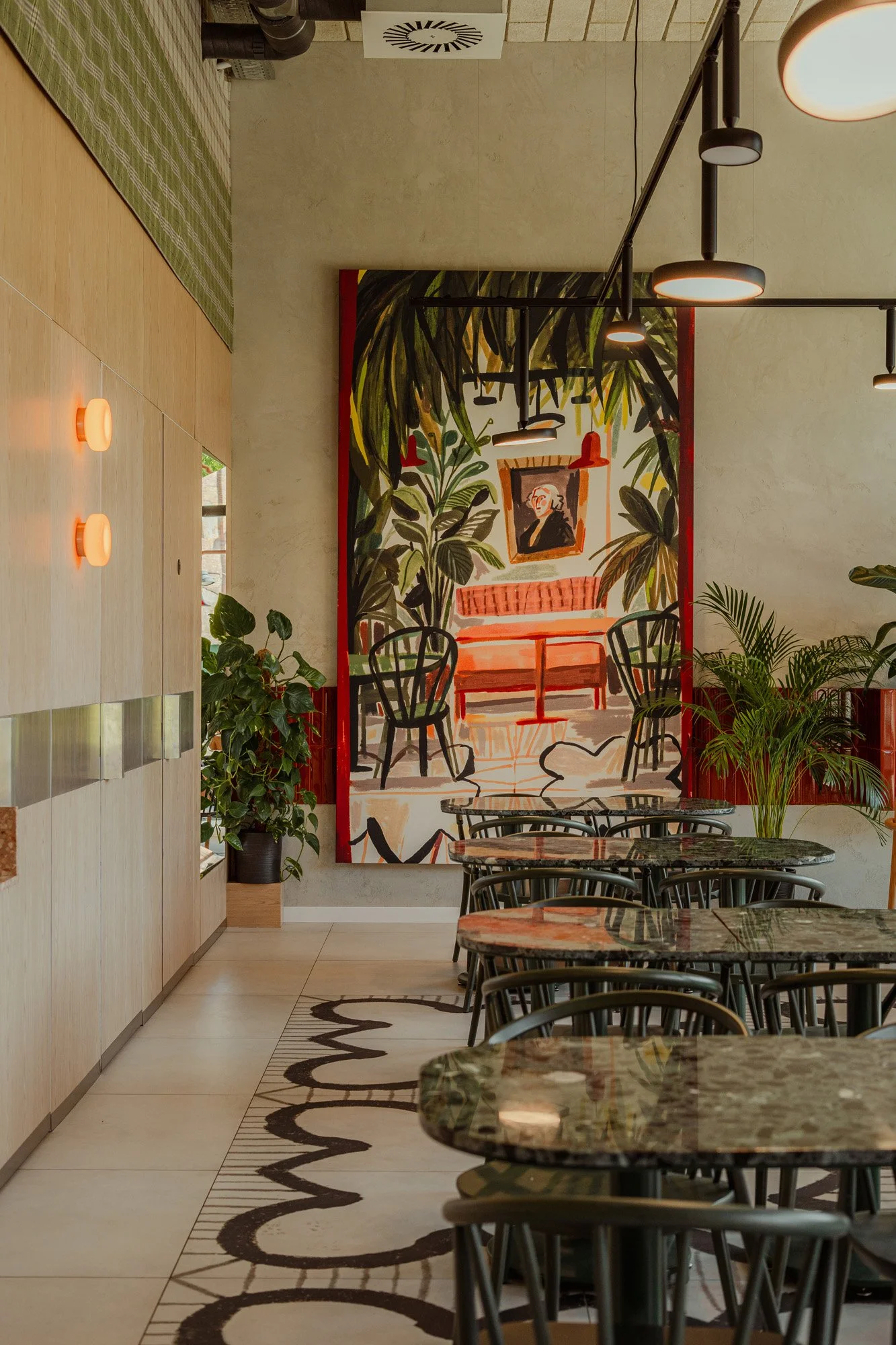 Interior of a modern cafe or restaurant with tables and chairs, a large colorful abstract painting on the wall, potted plants, and contemporary lighting fixtures.