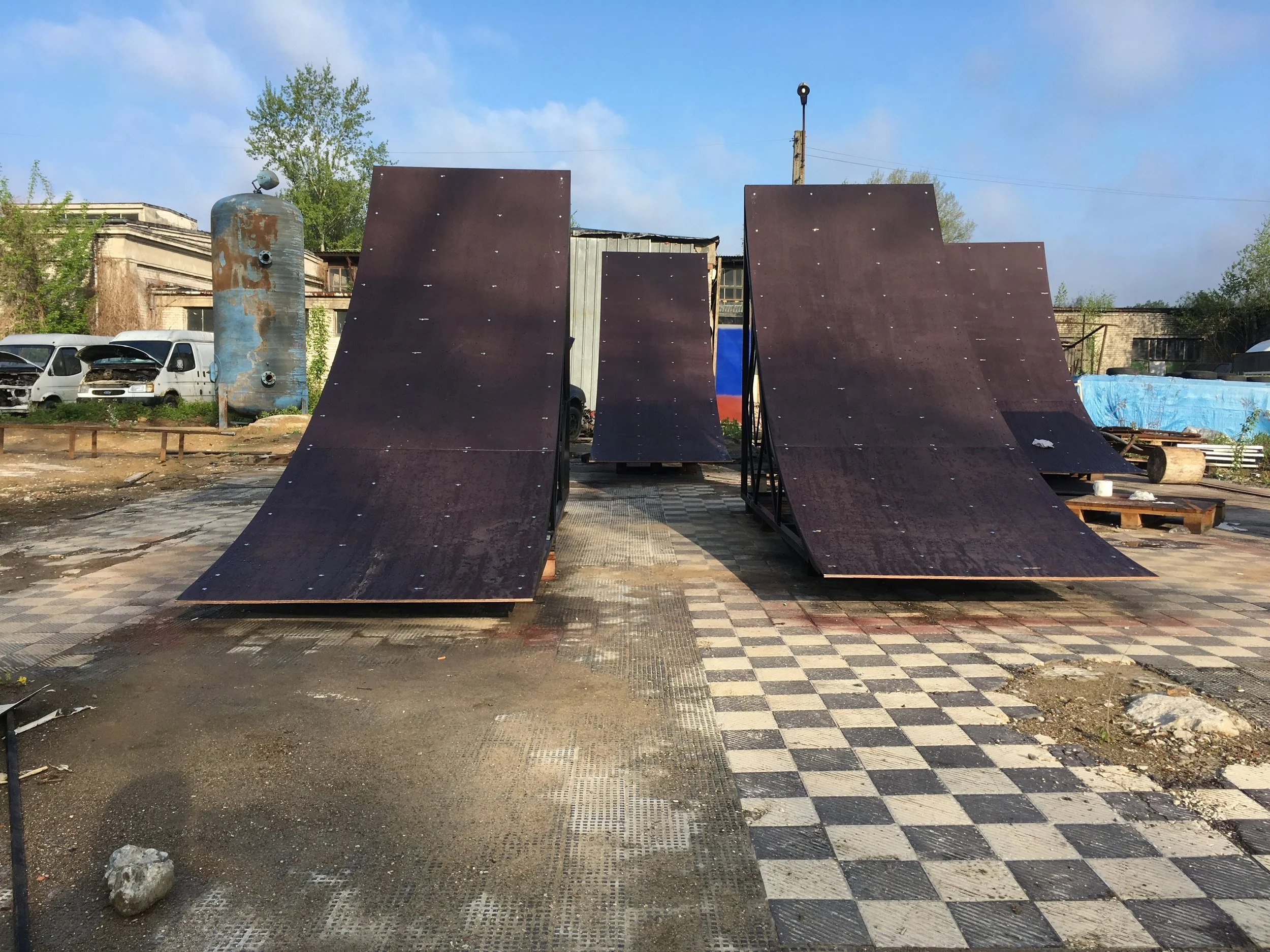 Skateboard ramps and quarter pipes set up on a checkered pavement outdoor area with industrial buildings in the background.