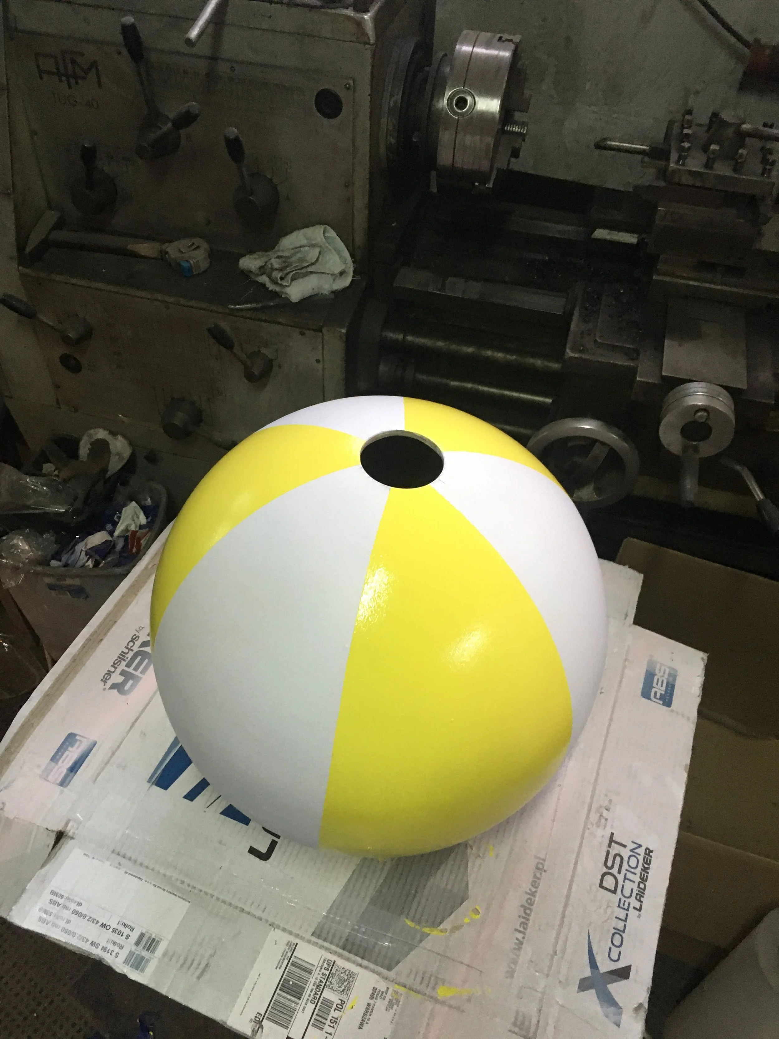 A large beach ball painted in yellow and white with a circular black hole on top, sitting on a cardboard box in a workshop with a metal lathe and other equipment in the background.