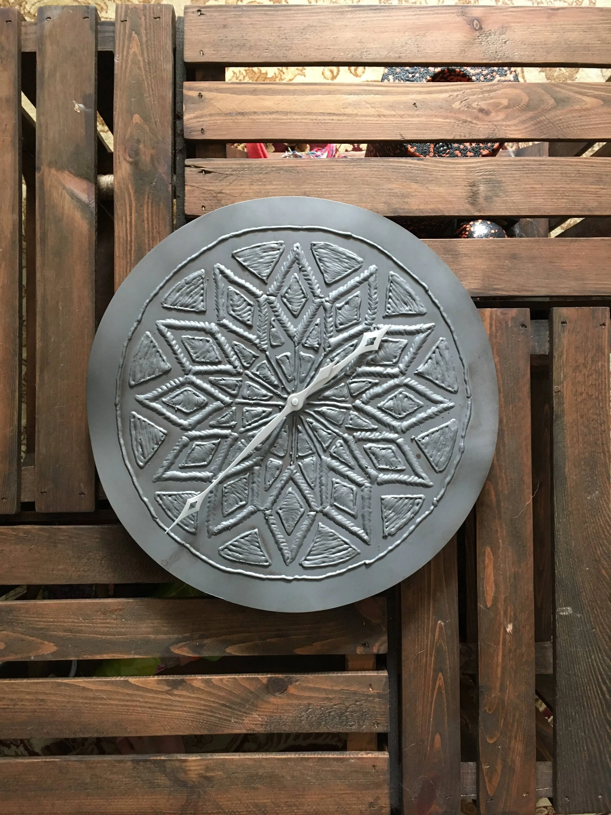 A round wall clock with a decorative geometric pattern on the face, showing the time as approximately 2:20. The clock is placed on a wooden slatted surface.