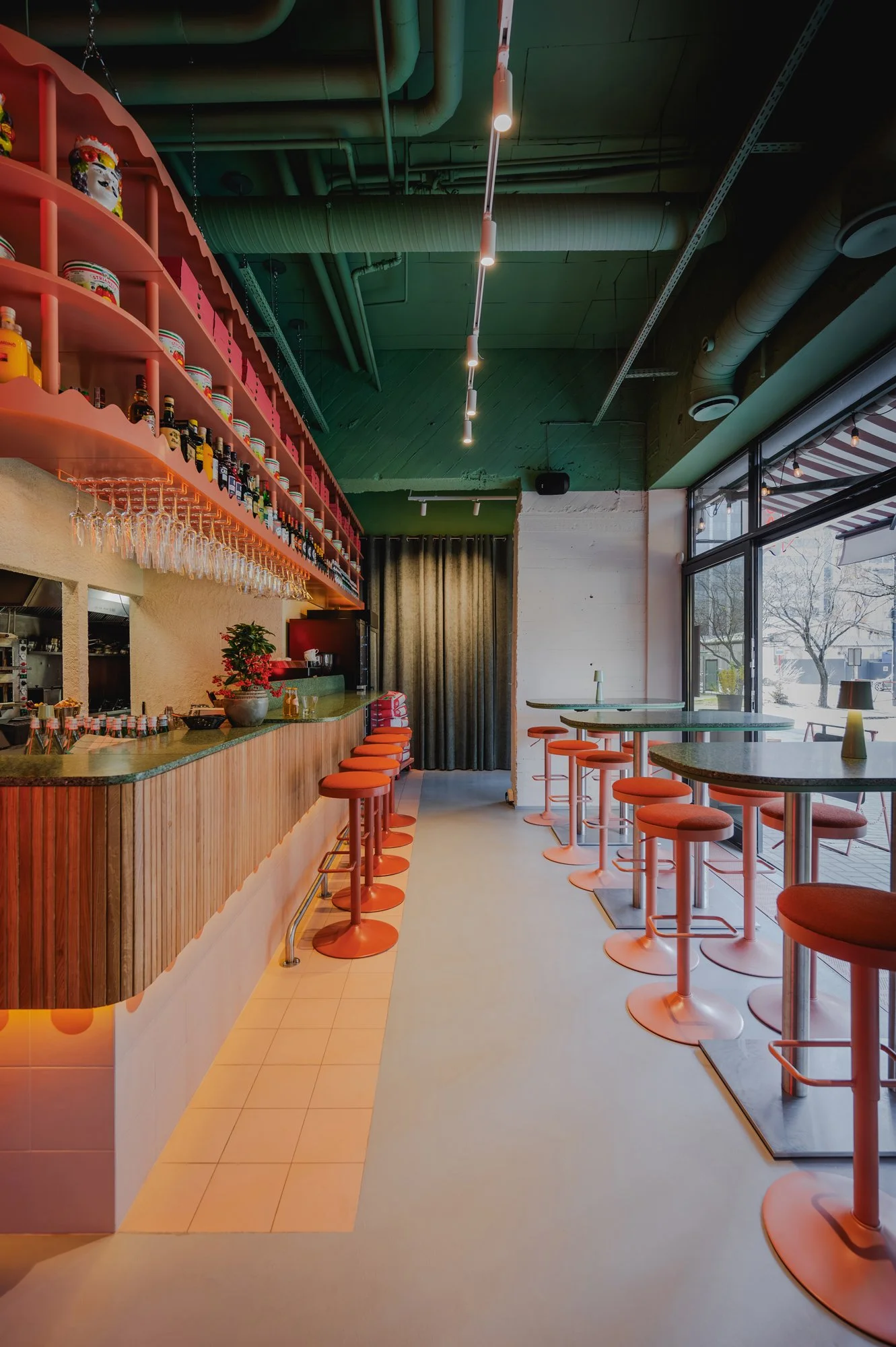 Interior of a modern restaurant or cafe with a bar counter on the left, seating with red stools and tables along the right, large windows at the front, and ceiling with exposed green pipes and lighting fixtures.
