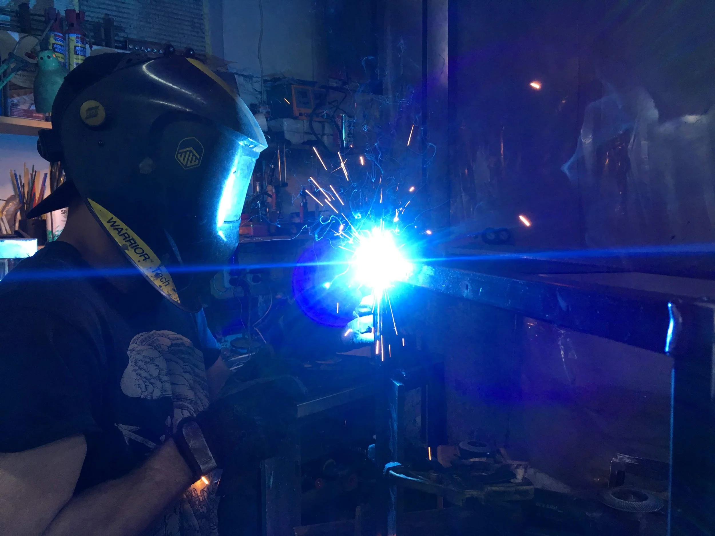 Person welding metal in a workshop, wearing a protective welding helmet and gloves, with a bright blue welding sparking.