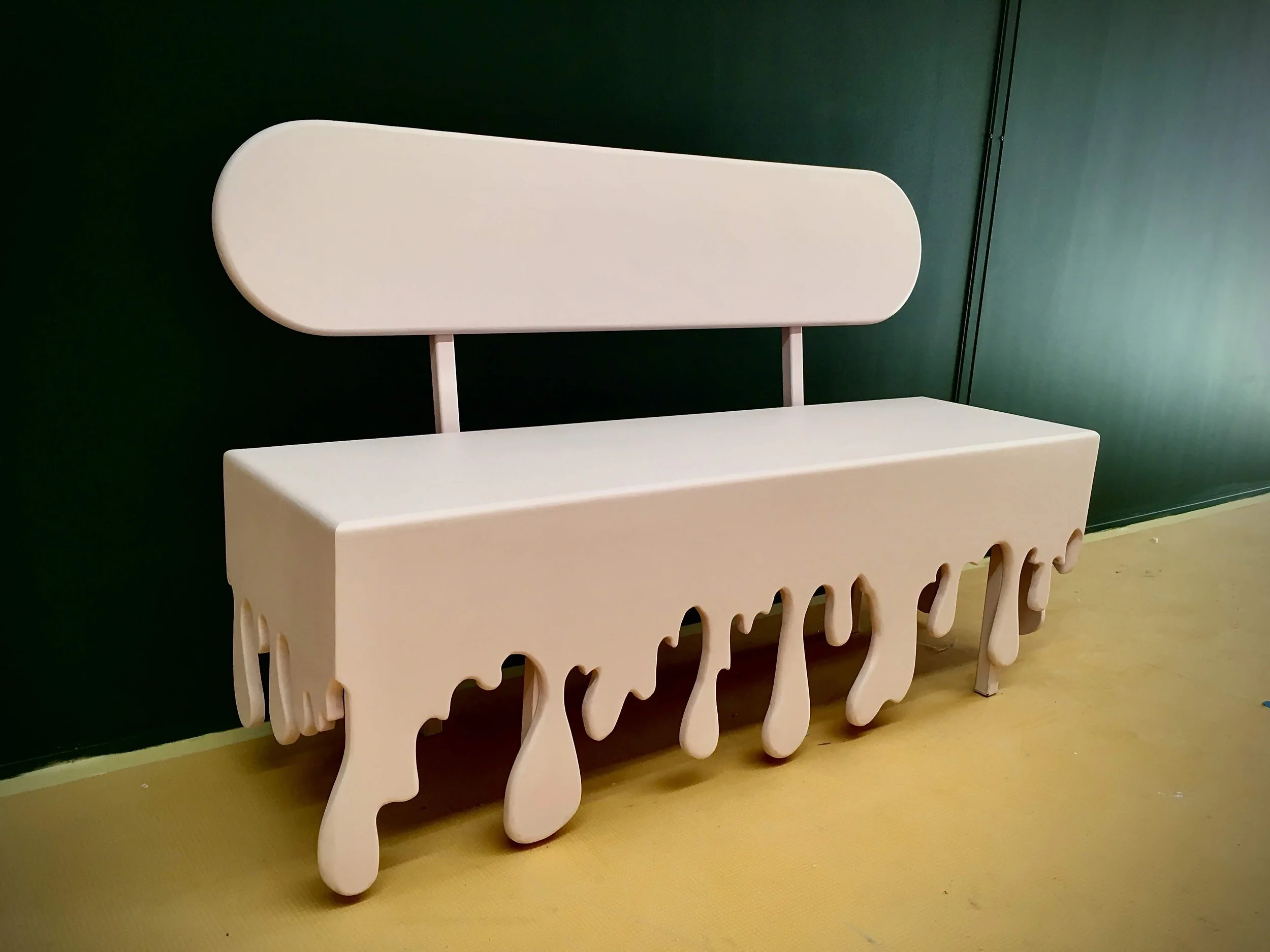 A white bench with a backrest, featuring a design that resembles dripping paint or melting wax, placed against a dark green wall with a yellow floor.