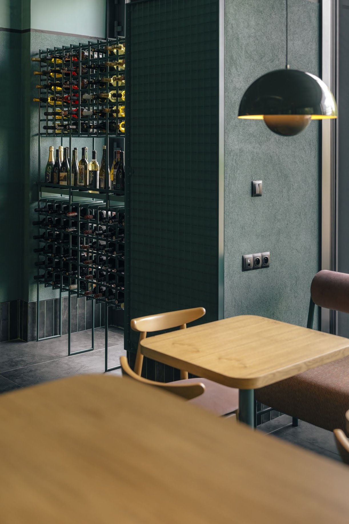 A wine storage area with bottles on horizontal racks, and some bottles on a lower shelf, in a modern interior with green walls, wooden and upholstered furniture, and a black and gold hanging light.
