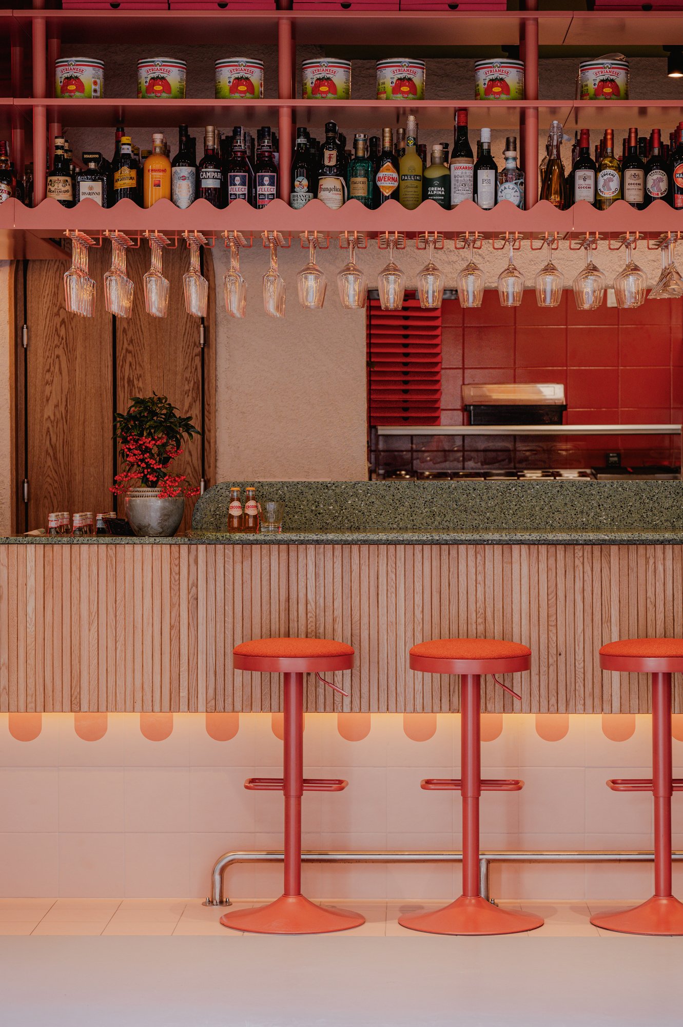 A bar with three orange barstools in front of a wooden counter with a green speckled surface. Behind the counter, there is a pink shelving unit holding bottles of various liquors and a row of upside-down wine glasses hanging. The background features 