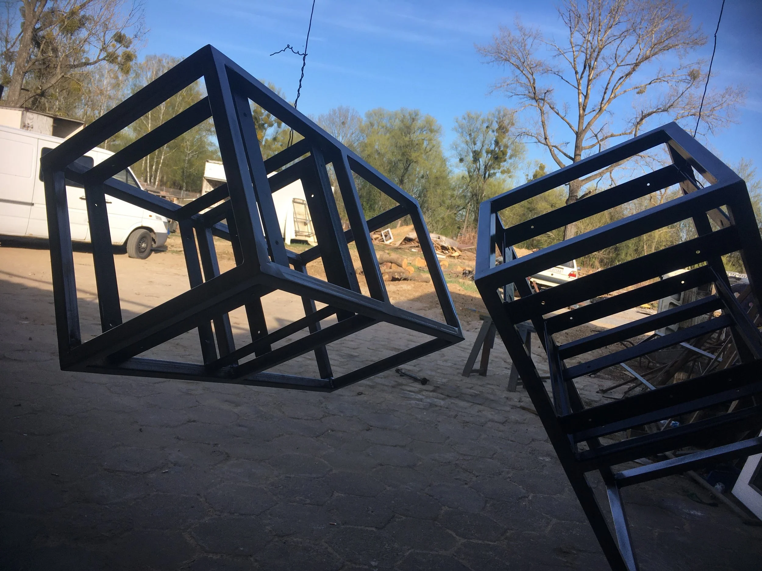Two black metal cube-shaped frames are suspended outdoors, with one tilted and the other upright. In the background, there are trees with bare branches, vehicles, and construction material on a dirt ground.