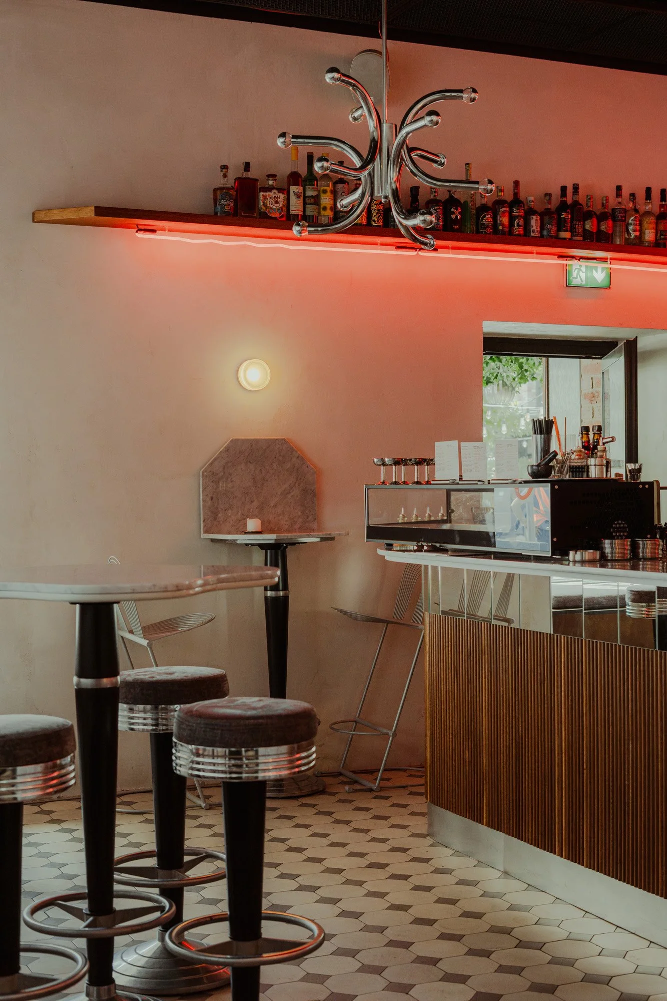 Interior of a modern bar with a wooden bar counter, bar stools, bottles on a shelf, and a unique chrome chandelier, with pink neon lighting and hexagonal floor tiles.