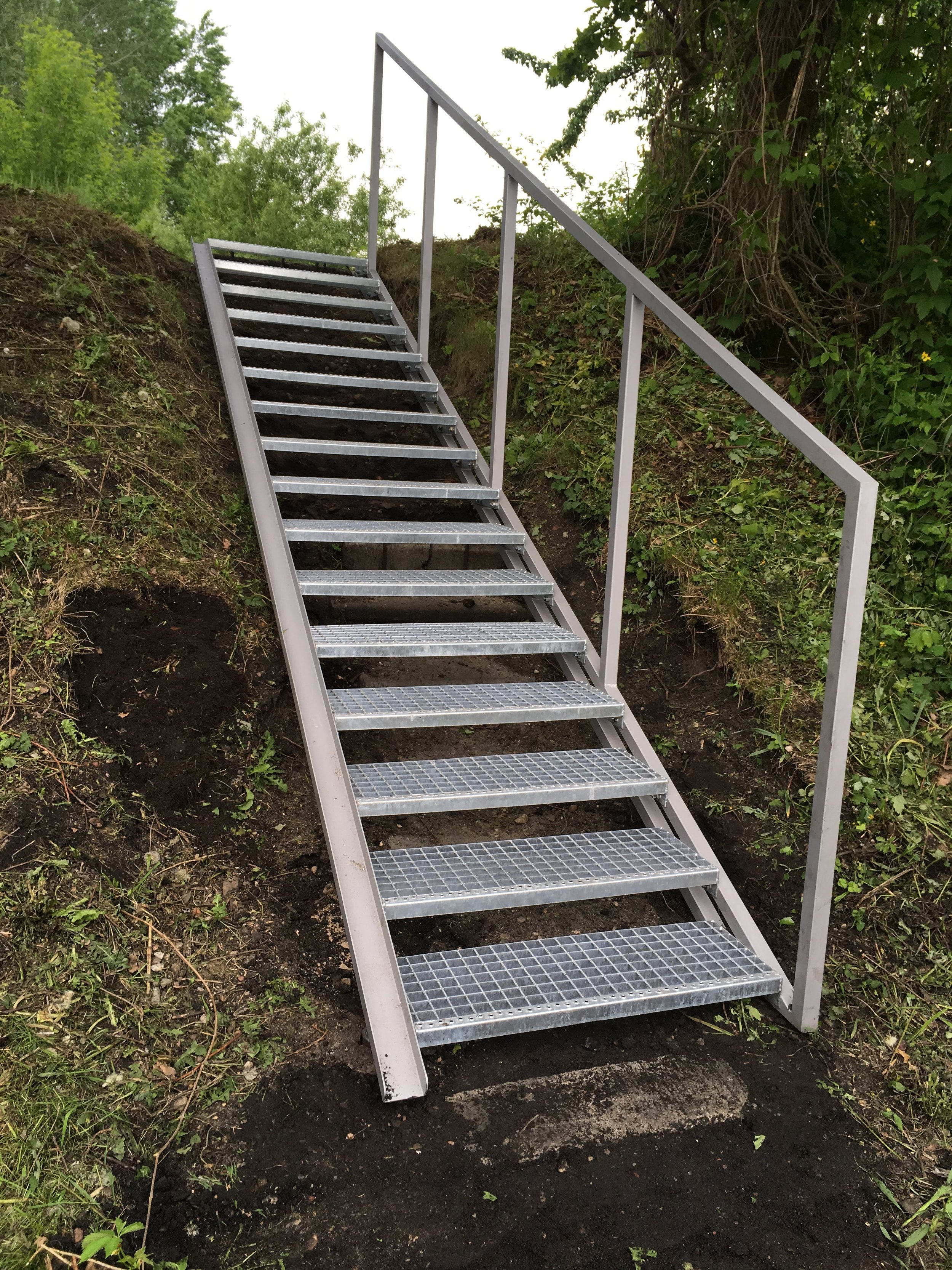 Metal staircase installed outdoors on a sloped dirt path with green bushes and trees in the background.