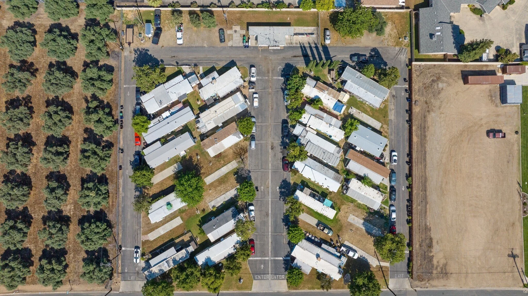 An aerial view of a small neighborhood with narrow streets, mobile homes, a parking lot, and a wooded area on the left.
