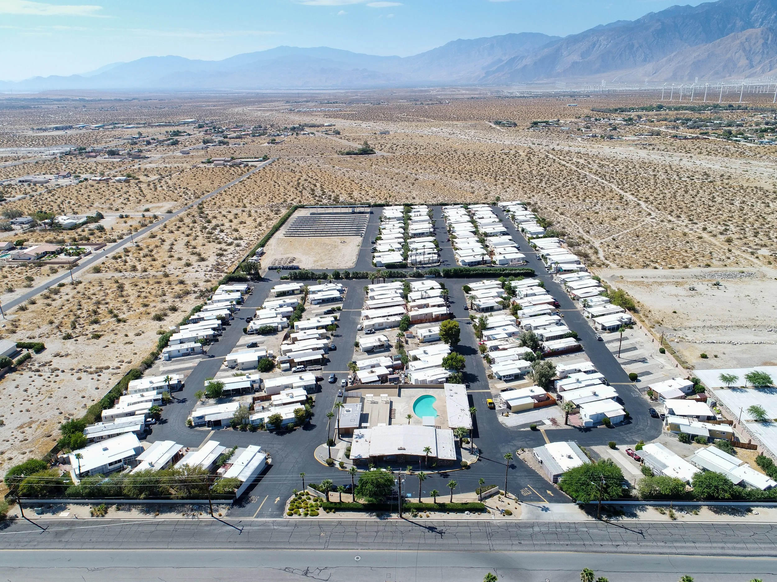 Aerial view of Park West mobile home park with rows of white trailers, paved roads, a central pool, and surrounding desert landscape. Mountains are visible in the background under a clear blue sky.