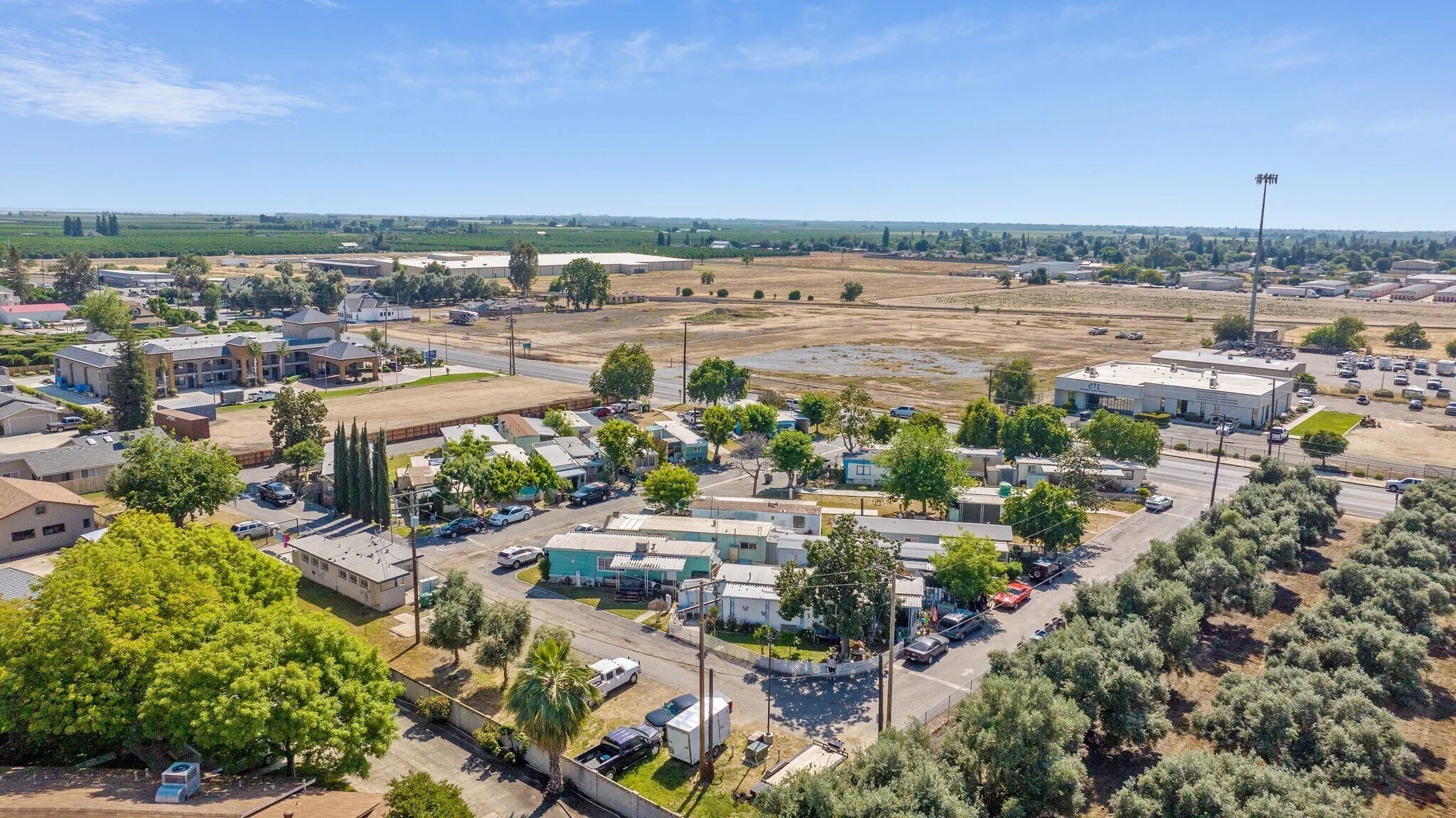 Aerial view of a kaweah mobile home community with rental houses, RVs, parked cars, a church, and open fields with farmland in the background under a clear blue sky.