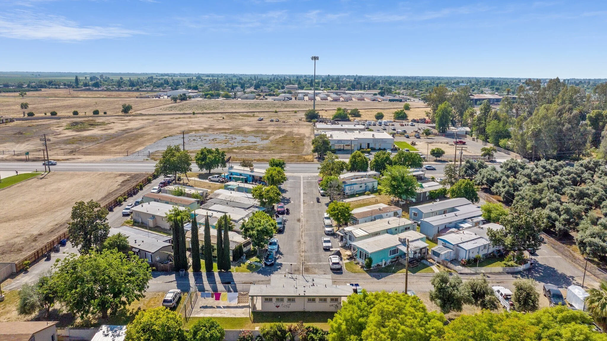 Aerial view of a Kaweah mobile home community with RVs and mobile homes, trees, parking lot, and a main road, with open fields and industrial buildings in the background under a clear blue sky.