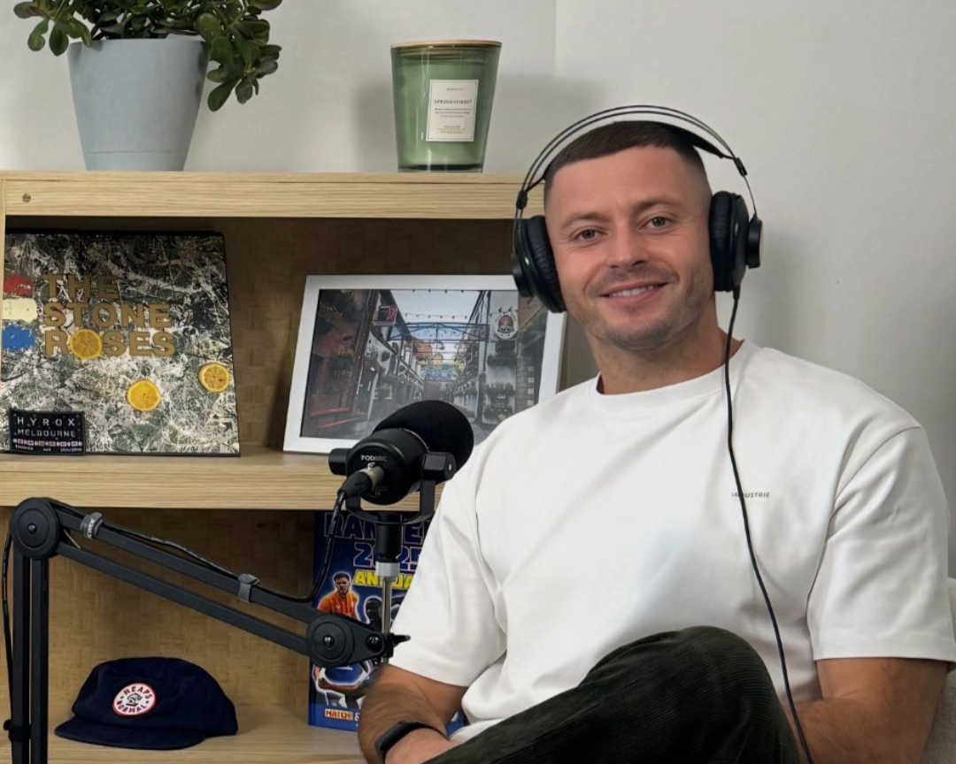 A man sitting in front of a microphone, wearing headphones, smiling, in a room with a wooden shelf behind him. The shelf holds a potted plant, a green candle, a vinyl record album, a framed photo, and a comic book.