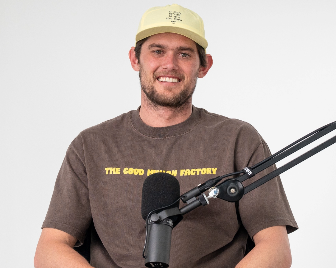 Man with a beard and short hair wearing a yellow hat and a dark brown T-shirt with yellow text, sitting in front of a microphone.