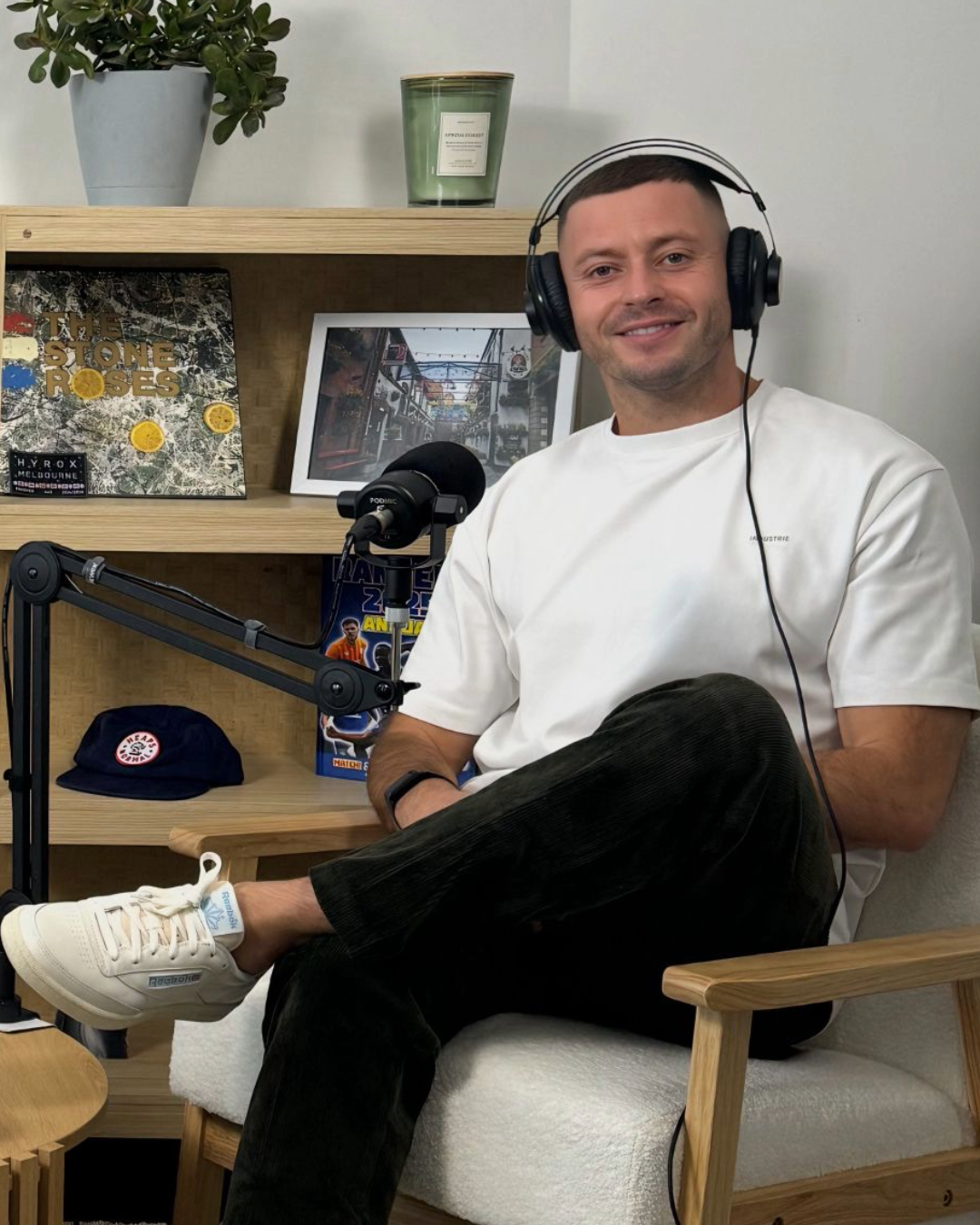 A man sitting in a chair with headphones, smiling at a microphone, in a room with a wooden shelf behind him holding a framed photo, vinyl records, and a cap. The man is wearing a white t-shirt, black pants, and white sneakers.