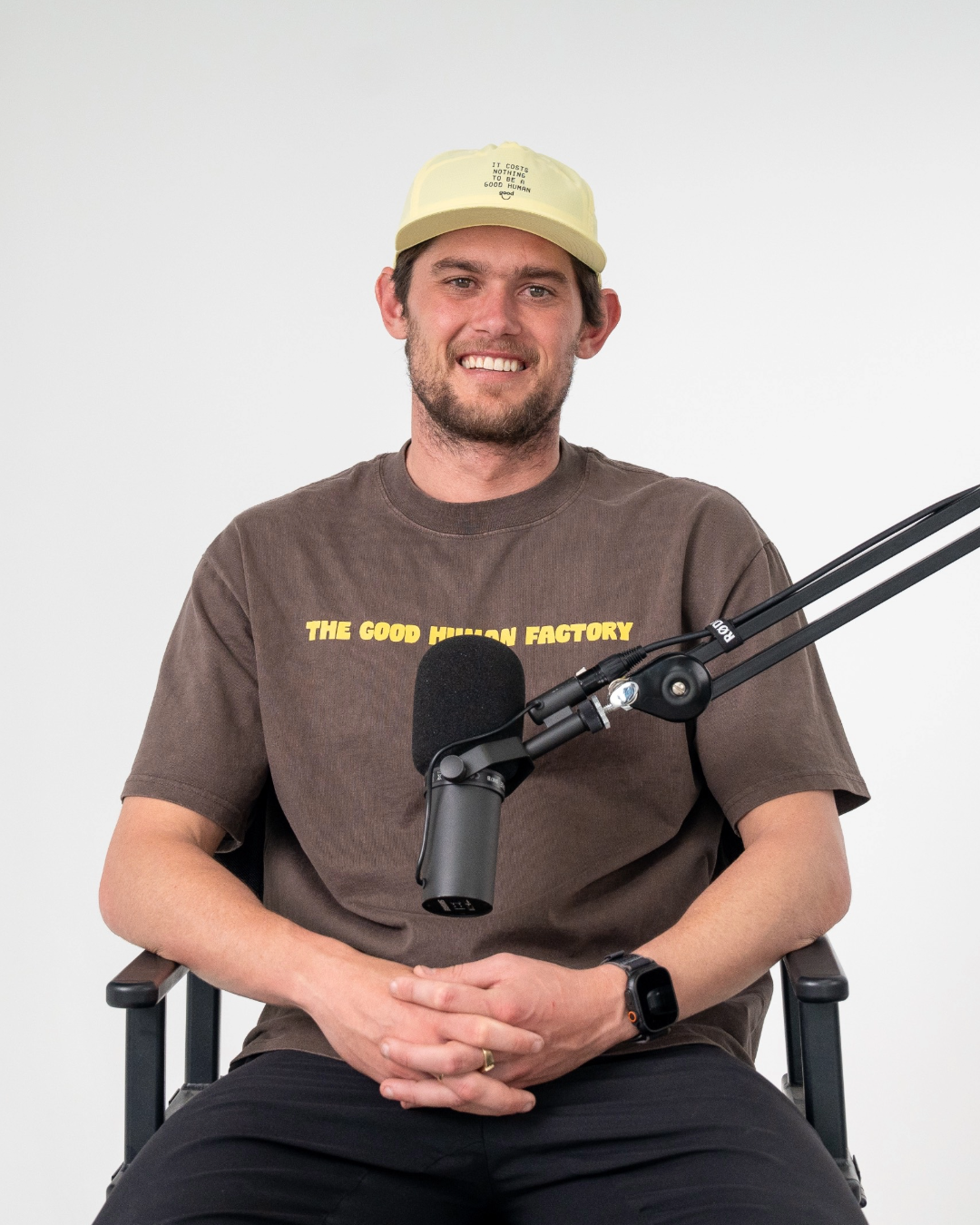 A man smiling, wearing a beige cap with text, a brown T-shirt with yellow text, sitting in front of a microphone, against a plain white background.