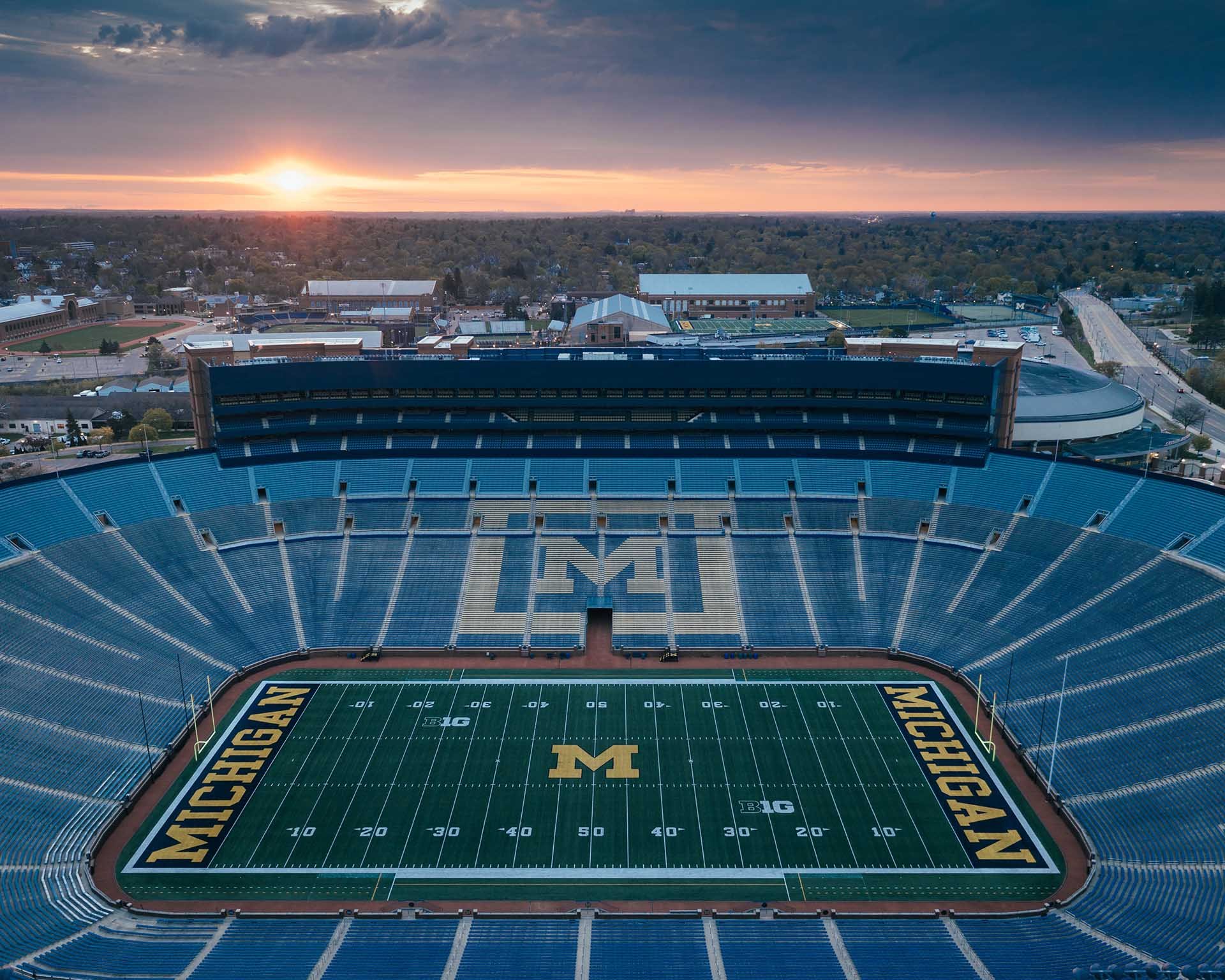 An empty football stadium at sunrise with a field marked with Michigan Wolverines branding and a large Michigan logo at the center. The background shows a cloudy sky and surrounding buildings.