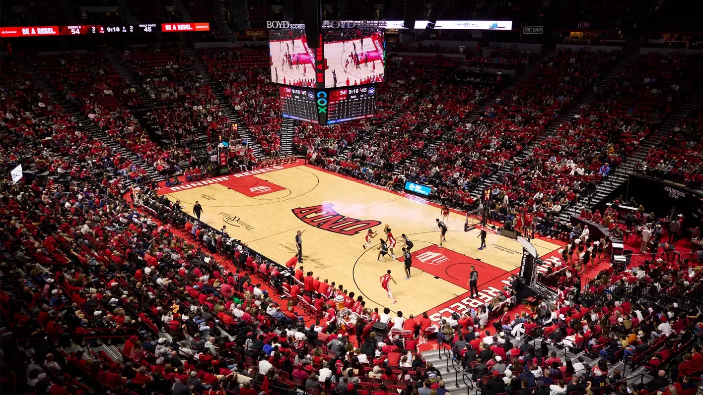 A basketball game in progress at the arena with players on the court, surrounded by a packed audience dressed mostly in red.
