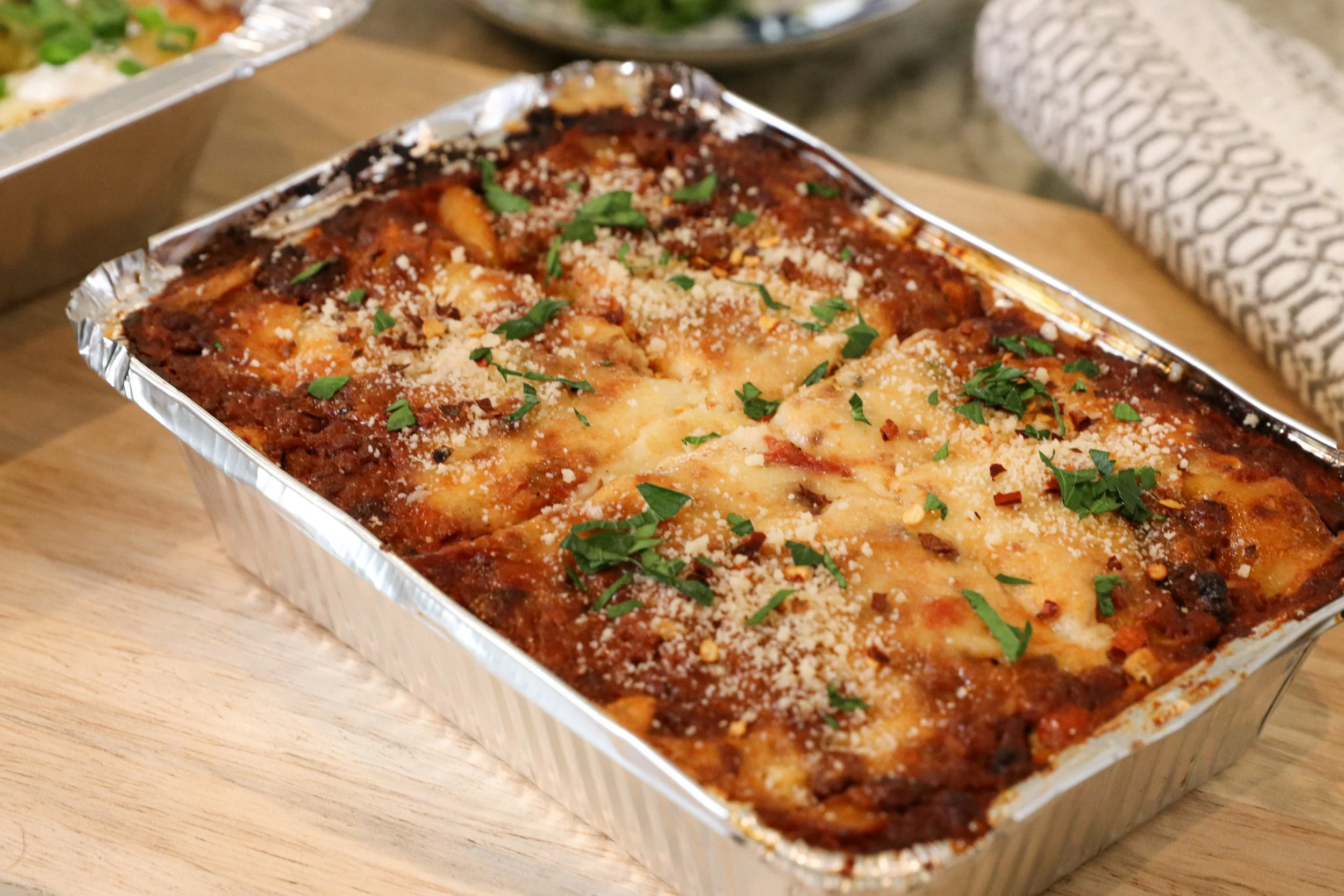 Baked lasagna in a aluminum tray garnished with parsley and grated cheese, sitting on a wooden surface.