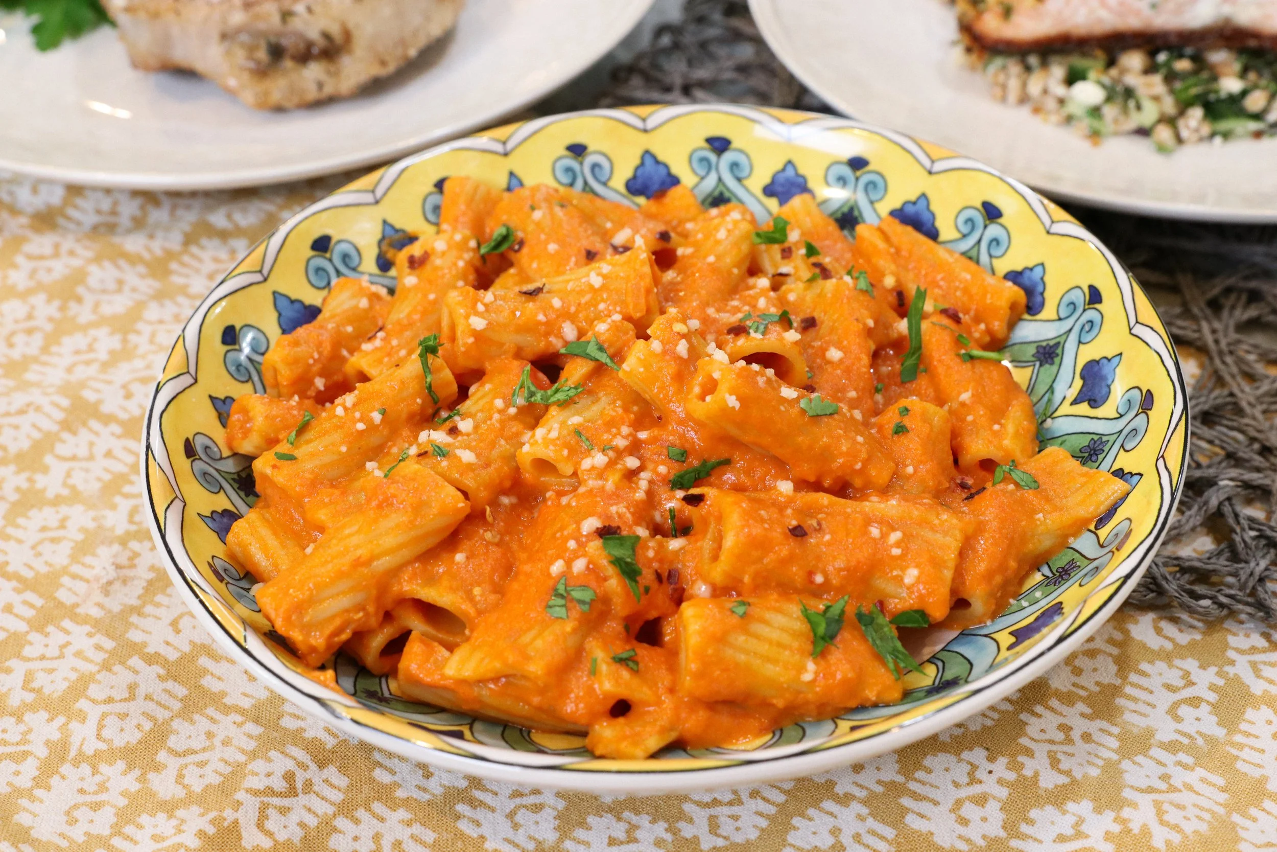 Plate of cooked pasta with orange sauce garnished with chopped parsley and grated cheese, on a yellow and blue patterned bowl with other dishes in the background.
