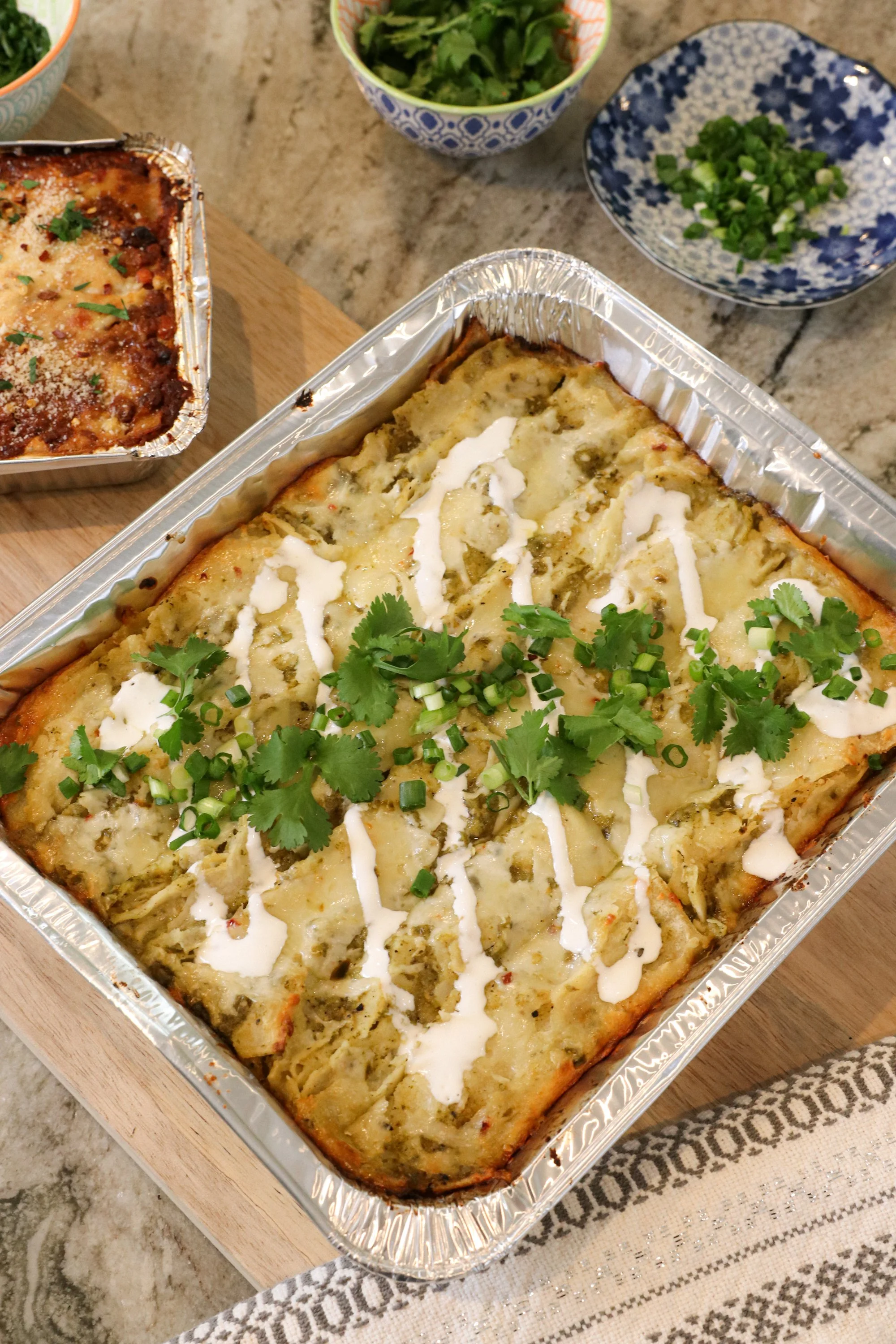 A tray of baked casserole topped with sour cream, chopped green onions, and cilantro, surrounded by bowls of chopped herbs and greens.