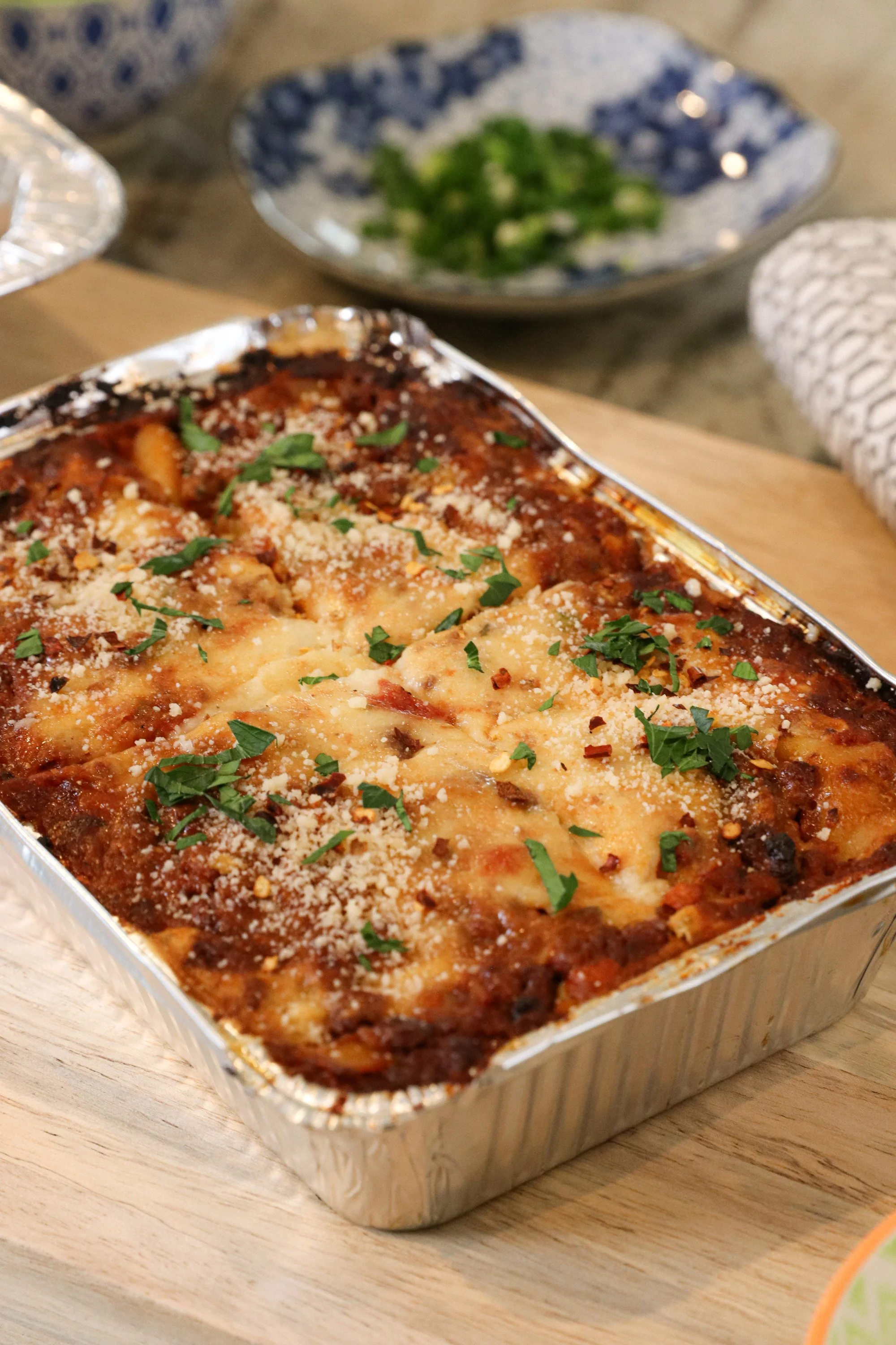 Baked lasagna in an aluminum tray topped with cheese and chopped herbs, on a wooden board.