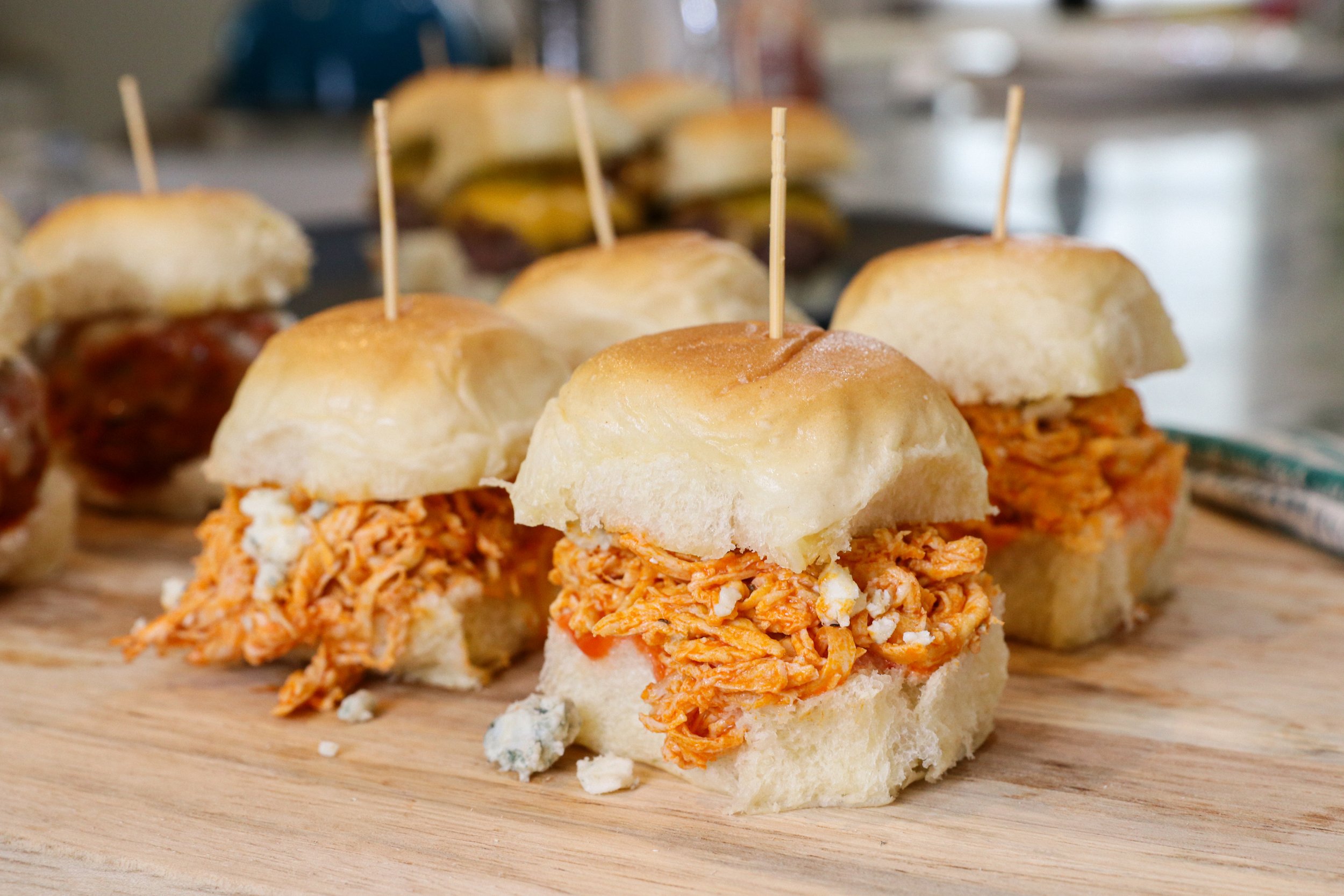 Close-up of chicken slider sandwiches on a wooden serving board with toothpicks, with other sliders blurred in the background.