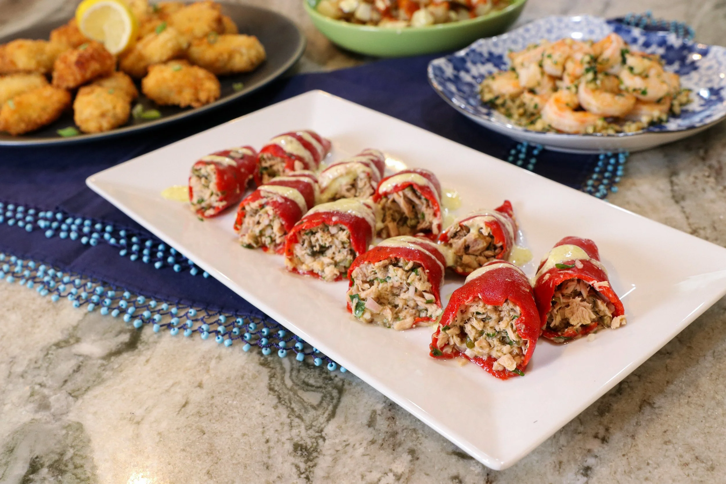 Close-up of a white rectangular plate with sliced stuffed red peppers filled with chopped vegetables and chicken, garnished with drizzled sauce. In the background, there are plates with fried chicken tenders with lemon slices, a bowl of shrimp in sau