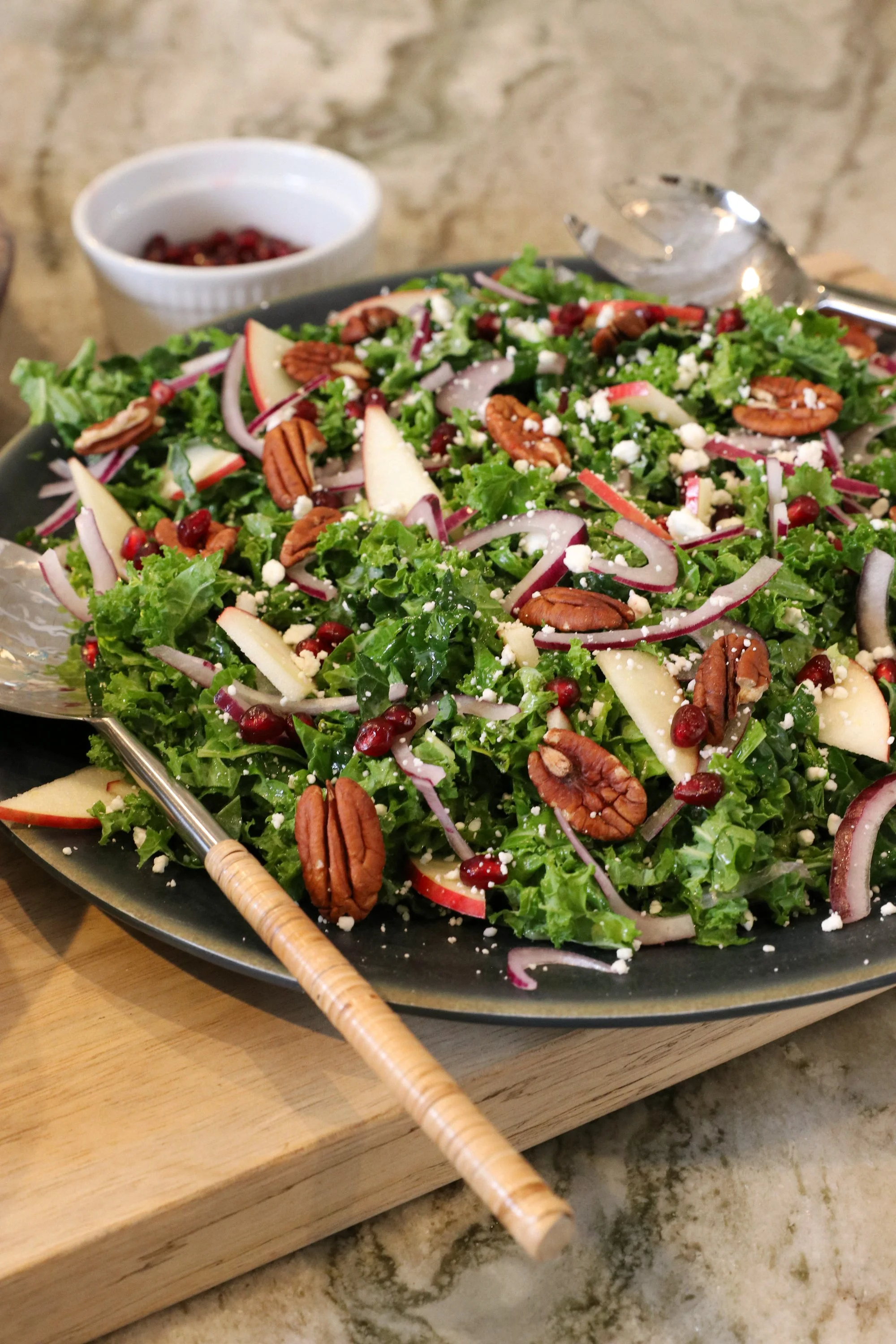 Fresh mixed salad with kale, apples, red onions, pecans, pomegranate seeds, feta cheese, and a bowl of pomegranate seeds in the background.